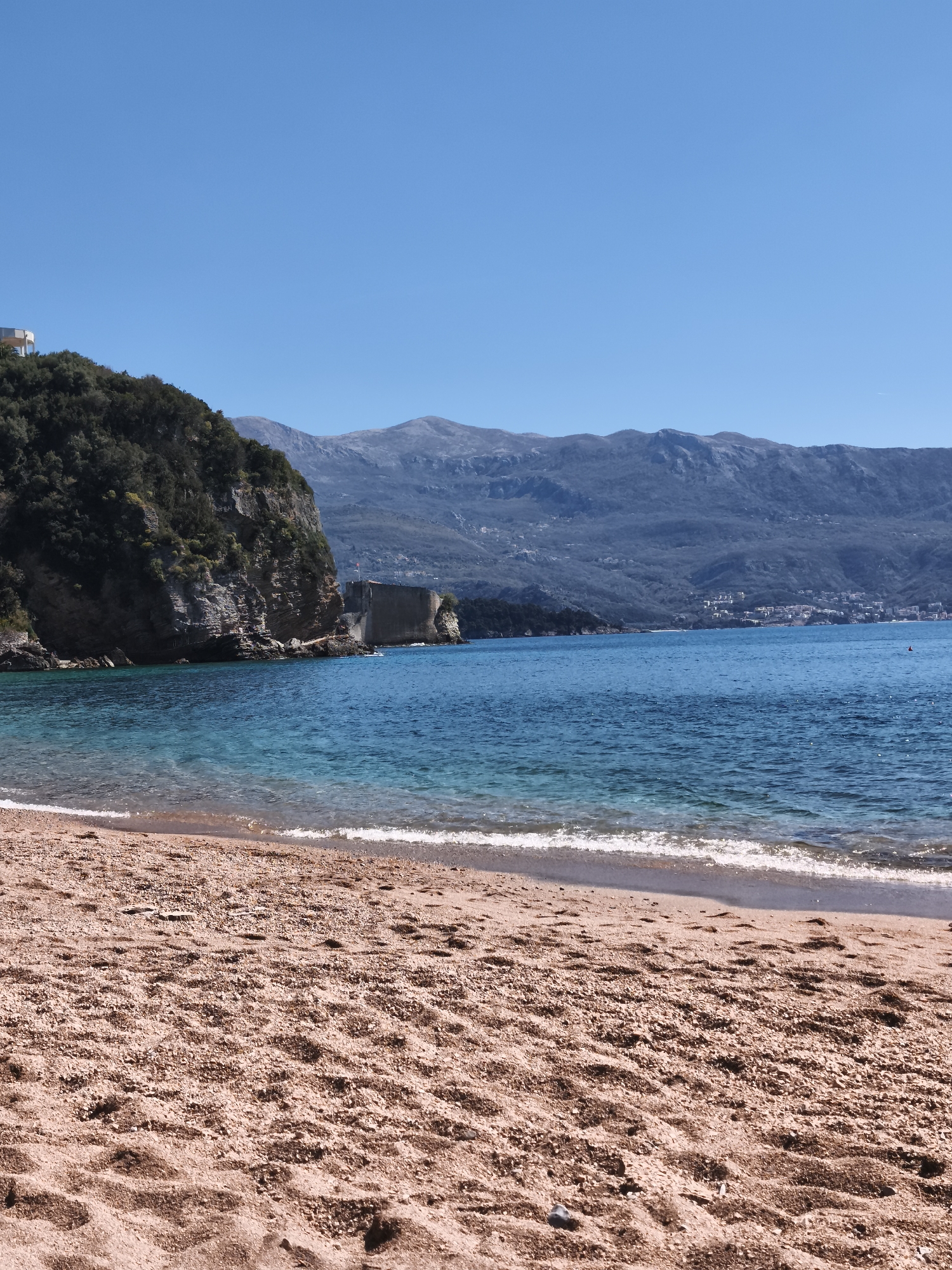 A coastal scene in Budva, Montenegro, features a sandy beach in the foreground. The blue Adriatic Sea meets the shore with small waves. To the left, a rocky cliff covered with trees extends into the sea, with a fortress on the rocks at the sea's edge. Mountains form the backdrop. The sky is clear and blue. No people are prominently visible in the scene.