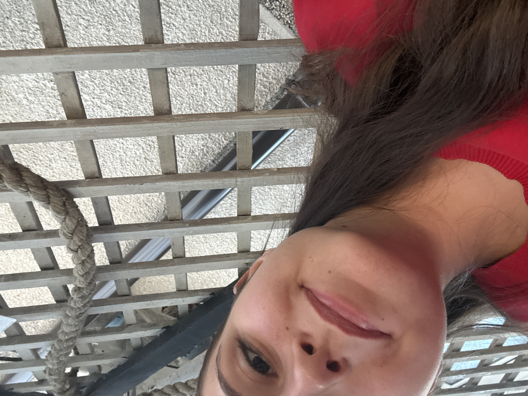 The image shows a person wearing a red shirt, with dark hair, in an outdoor setting. A light gray trellis structure is overhead. A thick rope is visible, possibly part of a nautical or decorative element. The background behind the trellis appears to be a textured surface. The location in Tsarevo, Bulgaria cannot be determined from this image.