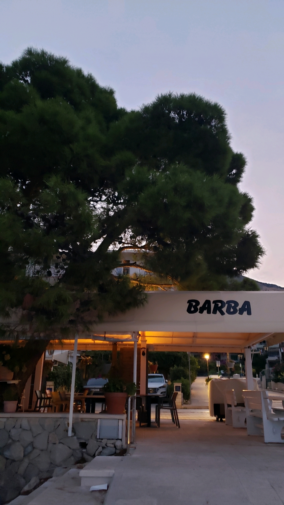 In Općina Tučepi, Croatia, at dusk, a waterfront establishment with an awning displays the word "BARBA" in black lettering. The awning is illuminated from below by warm, yellow lights, casting a glow on the seating area. Tables and chairs are arranged under the awning, some with visible place settings. To the left of the seating area, a large pine tree with dark green foliage extends into the frame, its branches partially obscuring a building with white balconies and windows. A stone wall borders the edge of the seating area. Beyond the awning, a paved pathway leads away from the establishment, with a white car parked further down the path and other vehicles and buildings visible in the distance. White benches are visible to the right of the pathway, some covered with tarps. The sky is a soft, pale purple, indicative of twilight.