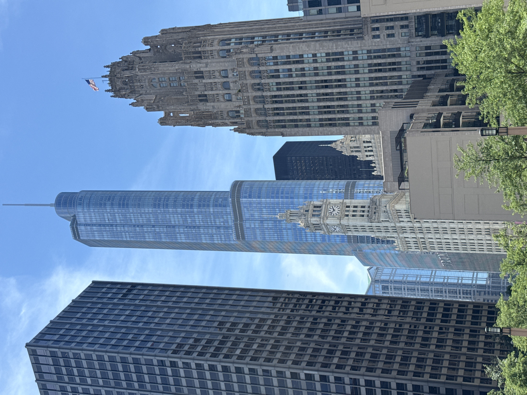 This image, taken in Chicago, Illinois, United States of America, depicts a cityscape dominated by several tall buildings under a bright, partly cloudy sky. On the far left is a dark, rectangular skyscraper with a grid-like pattern of windows. To its right and slightly behind is a very tall, slender skyscraper with a blue, reflective facade, topped with a spire. In front of this blue skyscraper, and to the right of the dark building, is a white building with a prominent clock tower. Further to the right is a Gothic-style skyscraper made of stone, featuring intricate architectural details and topped with an American flag. Several other modern buildings are visible in the background and between the main structures. Lush green trees line the bottom of the image, with some parts of modern buildings with balconies interspersed. The scene appears to be a street-level view of a dense urban environment.