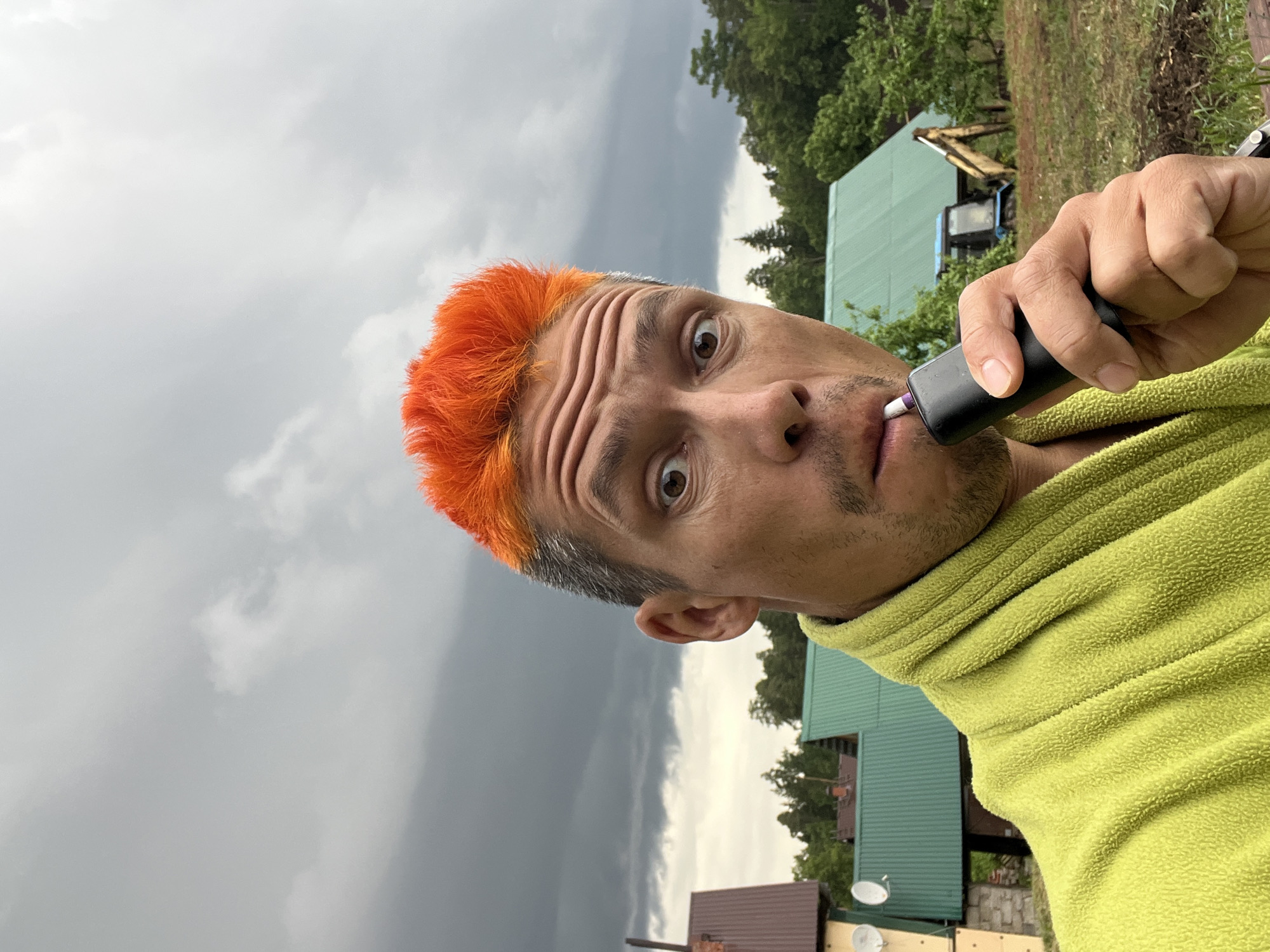 A man with orange hair is holding a black electric trimmer to his upper lip, presumably trimming his mustache. He is wearing a light green, fuzzy-textured hoodie. The background shows a stormy sky, greenery, and buildings, possibly in a rural or suburban area. A piece of heavy construction equipment, such as a backhoe, can be seen in the background. Given that the location is indicated to be Chaikovsky, Russia, the architecture of the houses appears consistent with Russian building styles.