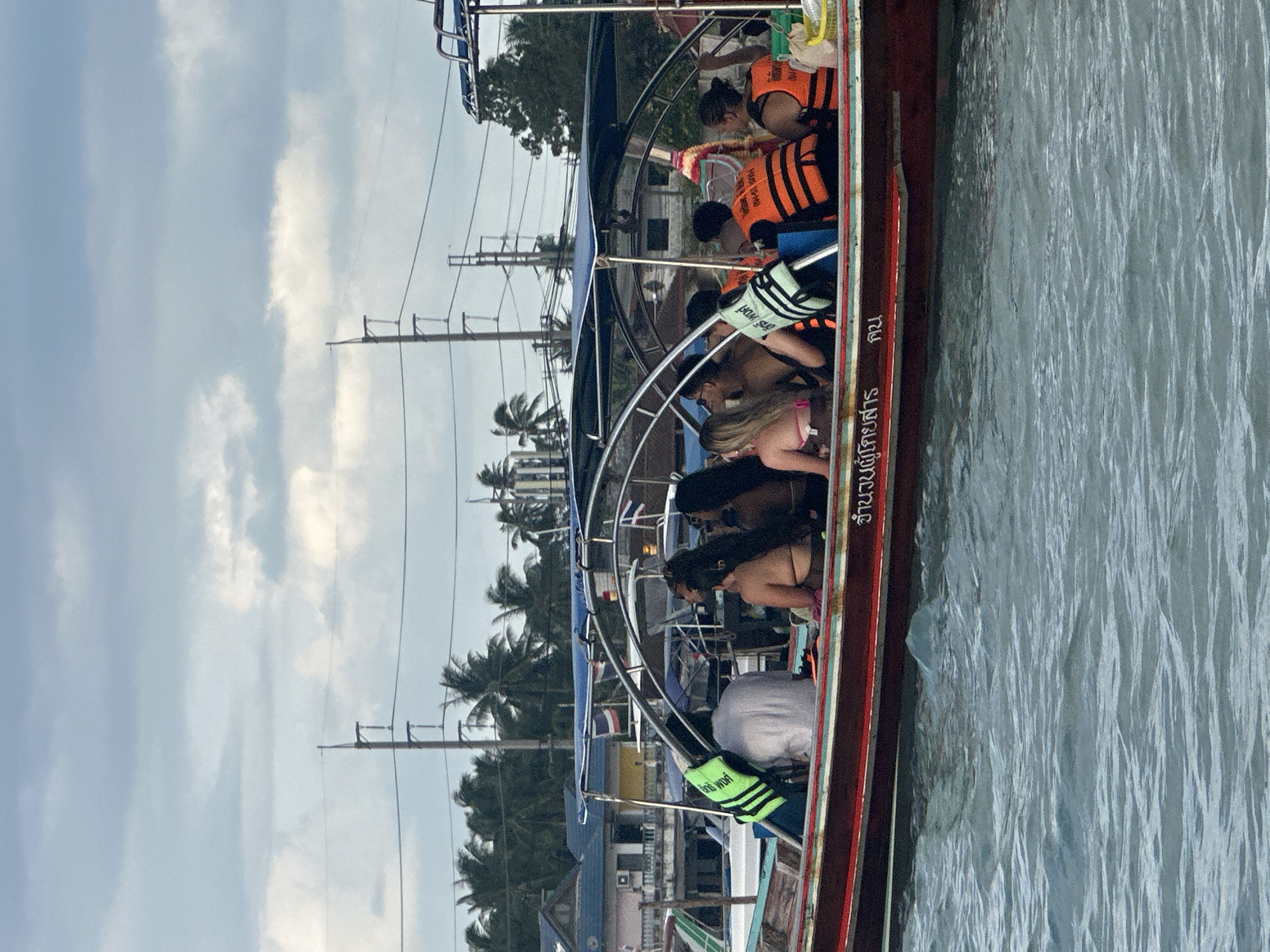 In Ko Samui District, Thailand, a long-tail boat is filled with passengers enjoying a boat trip on a body of water. The boat, painted red with a blue canopy and chrome railing, carries approximately eight individuals, some wearing bright orange life vests and one in a neon green vest. They are seated close together, facing forward and to the side. In the background, buildings with blue roofs and palm trees suggest a coastal town. Electrical poles and wires are visible against a cloudy sky, hinting at an urbanized area near the coastline. The water is choppy, indicating movement and possibly a breezy day.