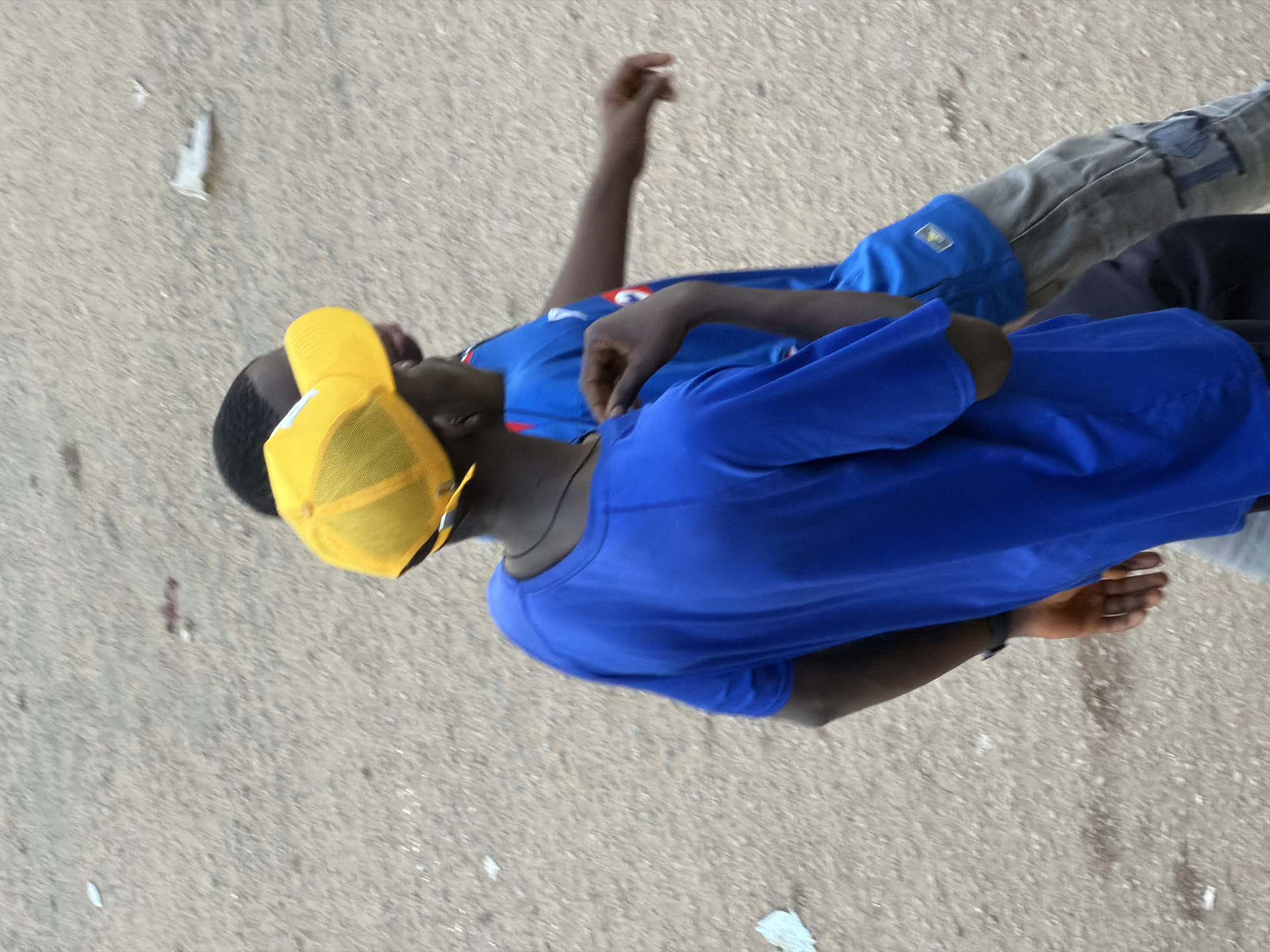 In Bukuru, Nigeria, two boys are walking on a dirt path. The boy in the foreground wears a bright blue t-shirt and a yellow baseball cap, his face obscured by the brim. The boy behind him is also wearing a blue shirt, partially visible. The path is composed of light brown dirt with scattered small pebbles and debris, indicating a dry, arid environment typical of some areas in Nigeria. The lighting suggests it is daytime.