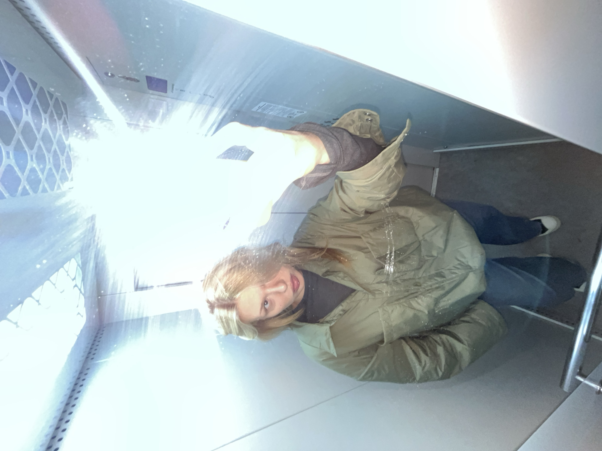 A single female individual is depicted in an enclosed, brightly illuminated space, consistent with an elevator car. The specific location within Chaikovsky, Russia, is not discernible from the visual evidence.
The individual, appearing to be a young adult female, has long, light blonde hair that exhibits a wet or damp appearance, with small water droplets visible on her hair and olive-green quilted jacket. She wears a dark long-sleeved shirt underneath the jacket, blue jeans, and white sneakers with dark soles. A small lip piercing is visible below her lower lip. Her gaze is directed straight ahead, towards the viewer.
Her right arm is raised and extended upwards, holding an activated, intense white light source, likely a flashlight or phone flash, which emits significant glare and lens flare, overexposing portions of the upper-right quadrant of the image. Numerous small, reflective water droplets are suspended within the path of this light.
The environment consists of light grey or metallic walls with vertical seams. The ceiling features a grid-like pattern, possibly a ventilation grate or light cover, and a row of small circular perforations along a horizontal beam. A metal handrail is visible in the lower-left foreground. On the right wall, a control panel for the enclosure is partially visible, showing multiple buttons and a distinct rectangular purple element. The floor is a darker, textured surface.