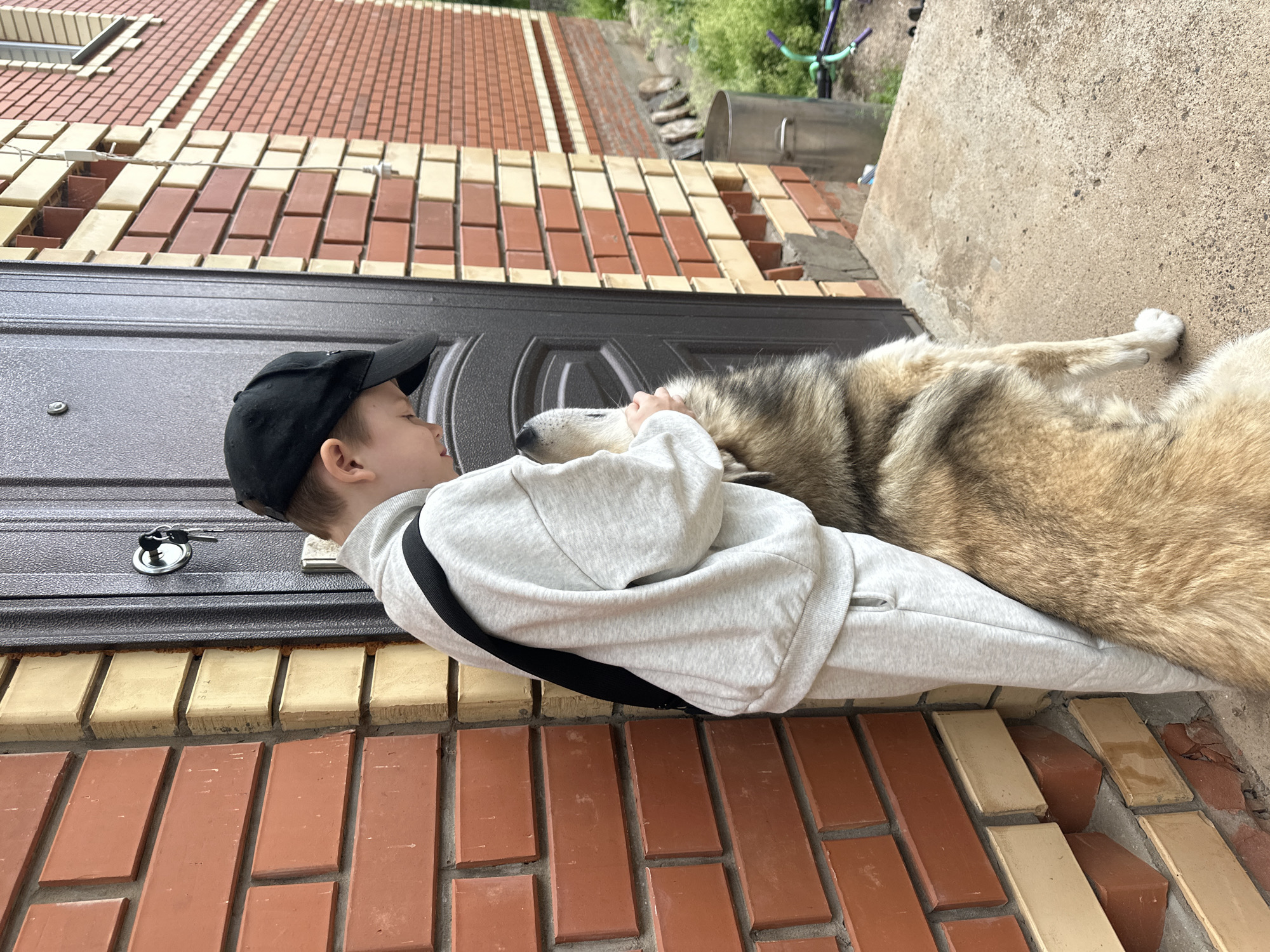 In Ilyinsky, Russia, a young boy wearing a grey tracksuit and a black baseball cap is standing next to a dark brown door. He has his arms around a large dog, which appears to be a husky or a similar breed with grey and brown fur. The boy is looking down at the dog, and the dog is looking up at the boy. They are standing on a concrete surface next to a brick wall. The brick wall is made of red and yellow bricks. To the right of the door, there is a view of a brick building and some greenery. A metal bin and some garden tools are also visible in the background. The scene appears to be outdoors, possibly in front of a residential building.