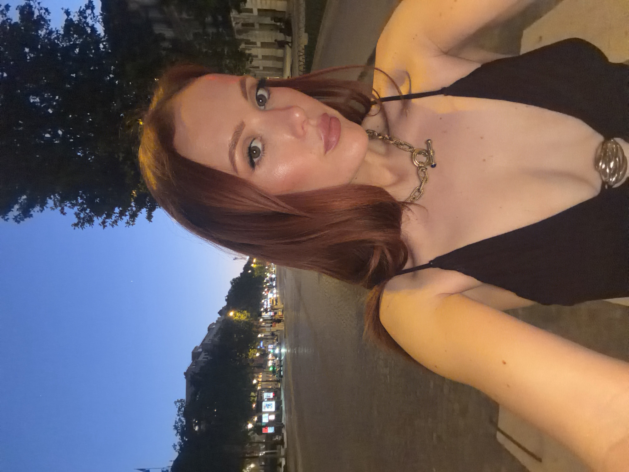 A woman with red hair is posing for a selfie at night. She is wearing a black tank top and a silver necklace. The location is the Champs-Élysées in Paris, France. Streetlights and a few buildings are visible behind her. The sky is a dark blue.