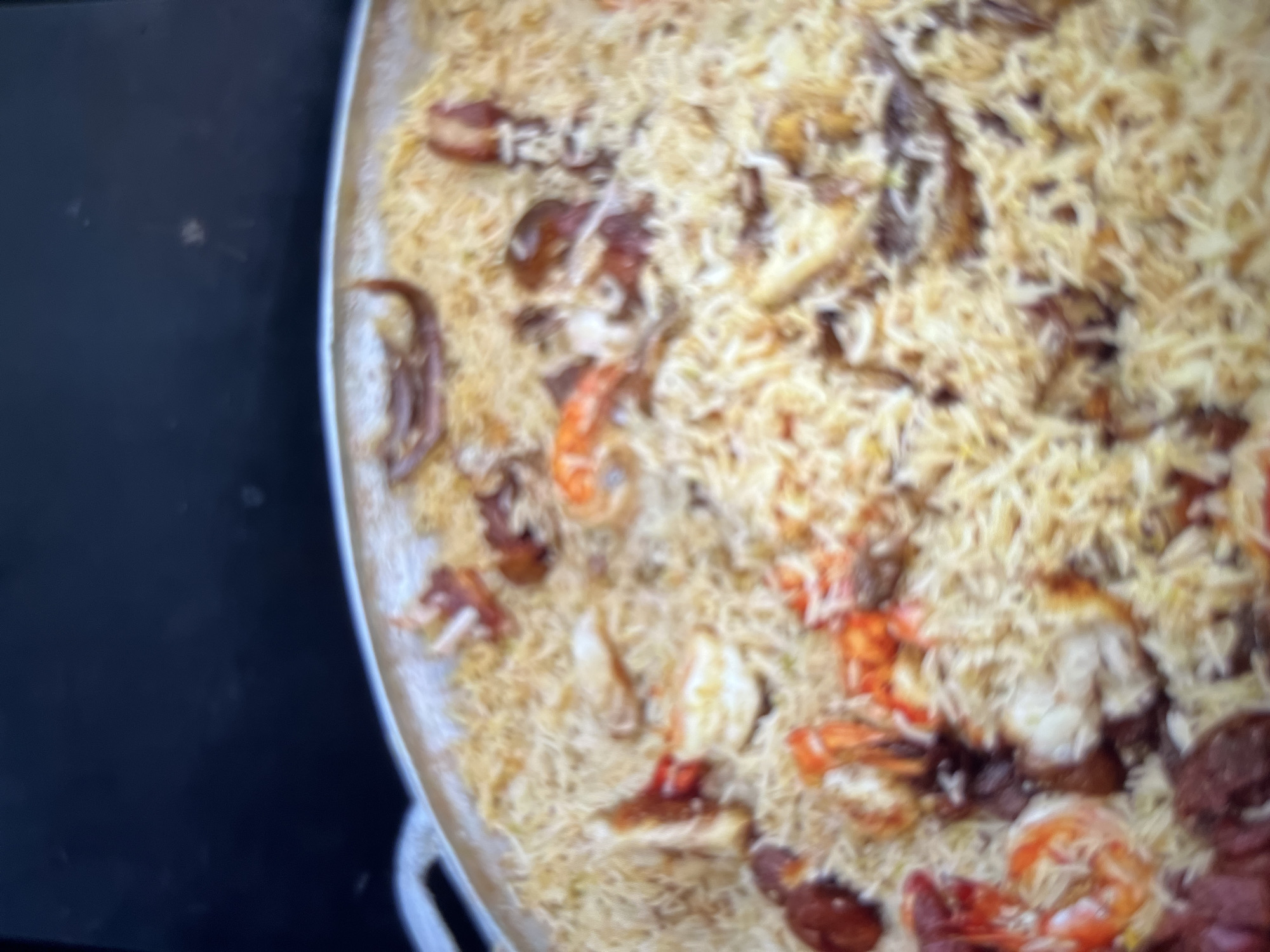 A close-up, high-angle shot shows a large, shallow metal pot filled with a rice dish mixed with seafood and what appear to be pieces of meat. The rice is a light golden color, and individual grains are visible. Scattered throughout the rice are pinkish-orange shrimp, some with their tails still attached, and dark brown pieces of meat. The pot is round and silver. The background is dark and out of focus, with a slight sheen suggesting it could be a countertop or a dark surface. The lighting is somewhat dim, creating soft shadows. There are no people visible in the image, nor any clear indication of the specific location within Abuja, Nigeria.