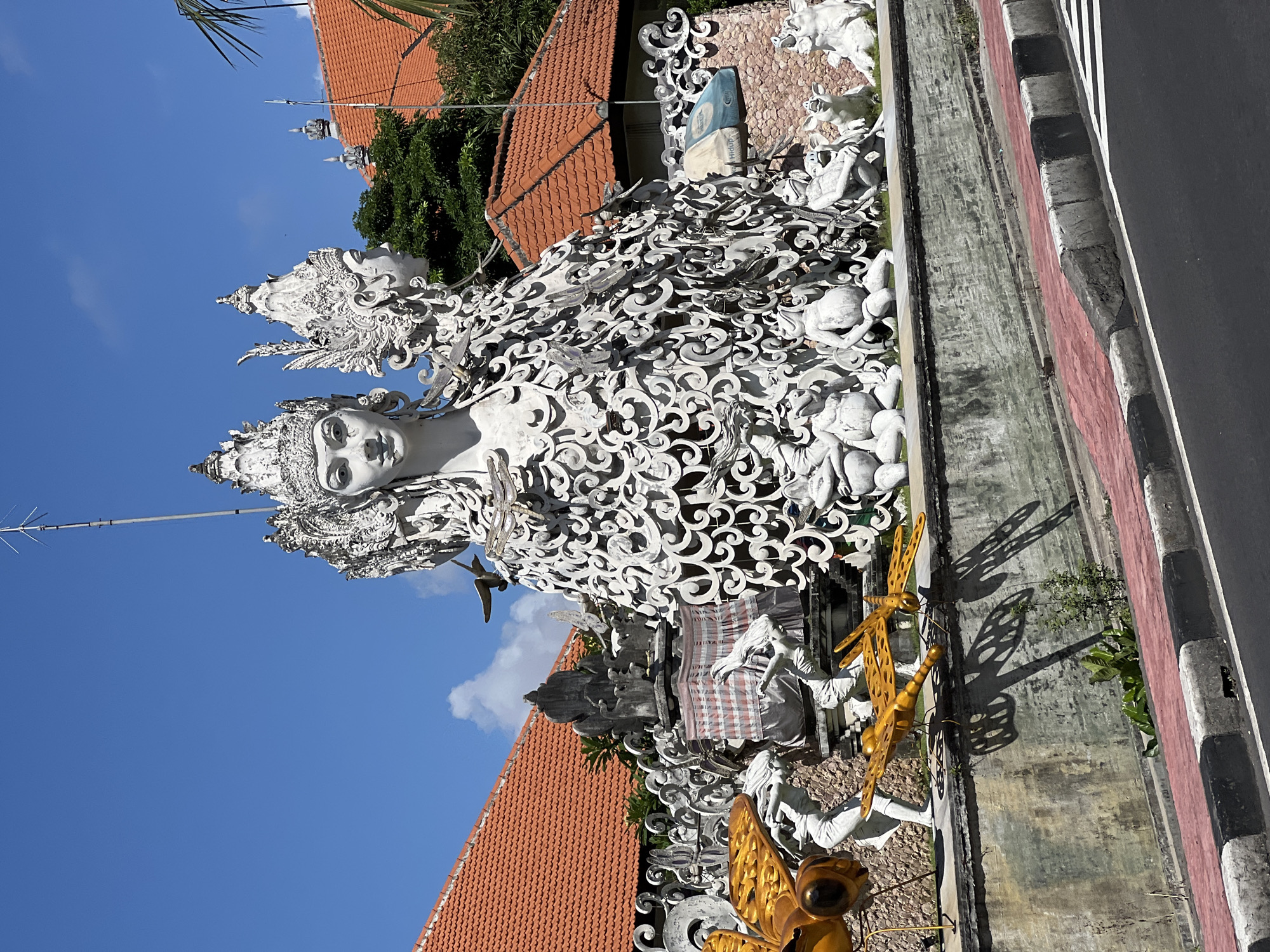 The image shows a large white statue with intricate details, possibly of a female deity or figure, adorned with complex carvings and patterns. Behind the statue are buildings with orange tiled roofs and greenery, set against a clear blue sky. In the foreground, there are two large metal dragonfly sculptures. The statue and sculptures appear to be part of a roadside display or public art installation. The location is likely Denpasar, Indonesia, based on the style of architecture and cultural elements present. The statue and sculptures are positioned on a concrete structure with a curb, with a road running alongside.