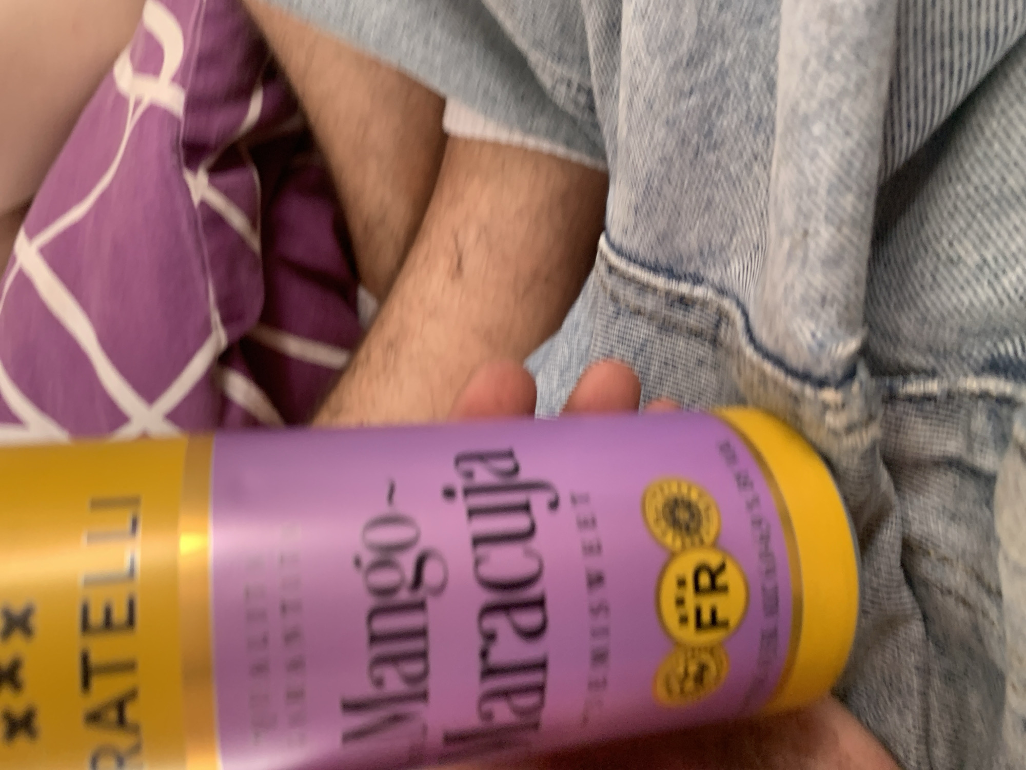 The image displays a close-up, slightly blurry view of a person holding a yellow and purple bottle. The bottle appears to be a beverage, with text that includes "FRATELLI," "Mango-Maracuja," and "FR." The person is wearing blue denim jeans. In the background, a portion of a purple pillow with a white pattern is visible, and parts of the person's arm and leg are also in frame. The overall setting suggests an indoor, casual environment, possibly a home. There is no identifiable information to place this scene within the city of Zhmerynka, Ukraine.