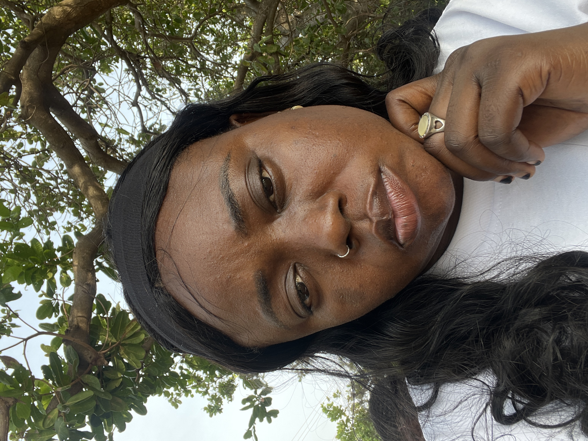 A medium shot of a Black woman lying down, wearing a white shirt and black headband. Her hair is long and black. She has a nose ring and is wearing a ring on her finger. The background consists of trees and the sky. The location cannot be determined with the provided information.