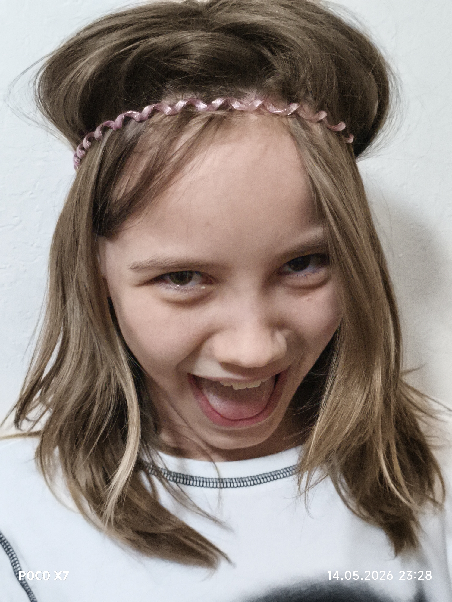 A close-up shot depicts a young girl with light brown hair and a pink headband. Her hair is styled up on top of her head. She is wearing a white shirt. The background is a plain white wall. The image date is 14.05.2026 23:28. The location is Unknown, Unknown.