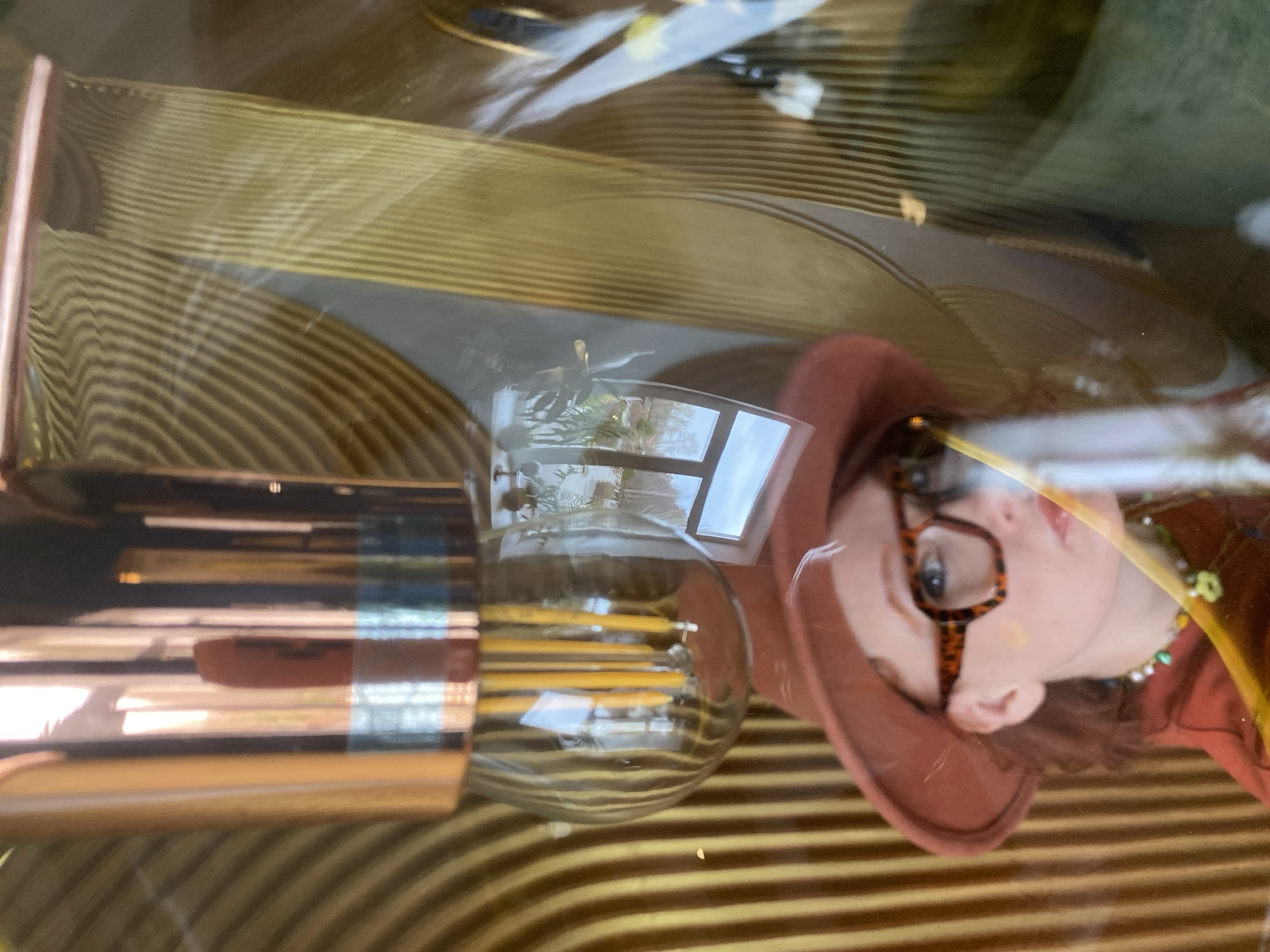 The image shows a display in a window, likely a shop window, with reflections overlaying the items on display. A woman wearing a hat and patterned eyeglasses is reflected in the glass. A cylindrical metal object is lying on its side, with a large light bulb next to it. The background is a striped pattern, possibly wallpaper or fabric. The reflection of a building and sky suggests an outdoor environment. The presence of indoor plants and daylight indicates the location is likely a commercial area in Yekaterinburg.