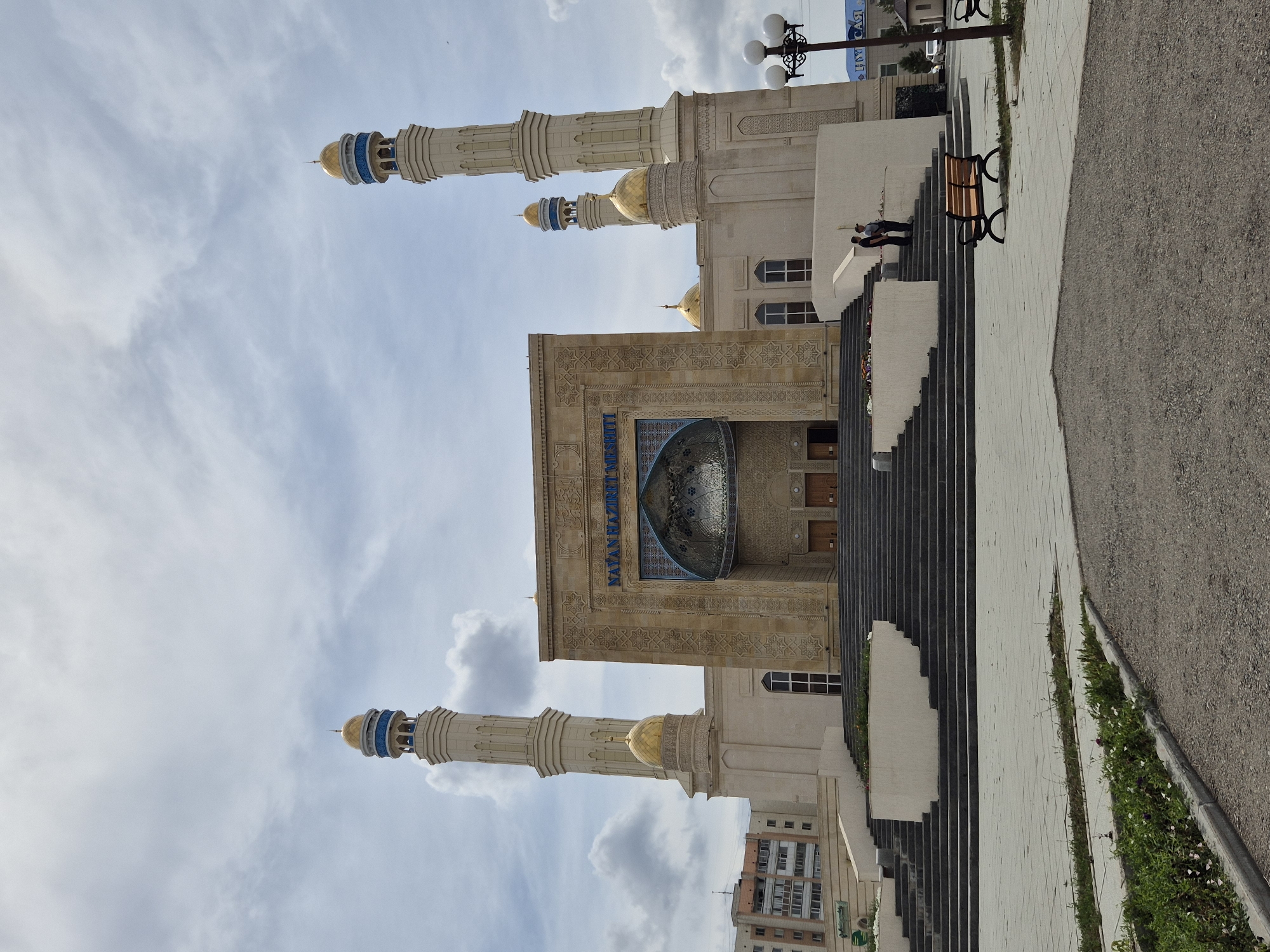 The image shows the Nur Astana Mosque in Kokshetau, Kazakhstan. The mosque is a prominent building with a beige facade, featuring two tall minarets topped with gold domes and blue accents. The central structure of the mosque has a large, ornate entrance and also a gold dome. A wide set of stairs leads up to the mosque's entrance from a paved plaza. Two men are standing on the stairs, facing the mosque. A park bench is visible in the foreground on the right side of the plaza. The sky is cloudy.