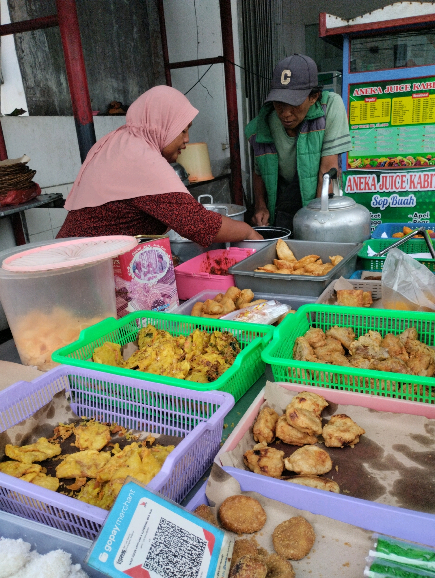 This image depicts an outdoor food stall in Bekasi, Indonesia. A woman wearing a pink hijab and a patterned red shirt is seen behind a counter, with her hands engaged in food preparation. To her right, a man wearing a grey cap and a green vest over a light green shirt is attending to the stall, leaning forward. In front of them, various food items are displayed in colorful plastic baskets and trays. These include fried snacks like tempeh and banana fritters, as well as what appears to be packaged biscuits. A juice menu board is visible in the background, advertising "ANEKA JUICE KABIT" and "Sop Buah". A prominent feature in the foreground is a GoPay QR code payment display, indicating the modern payment methods available at the stall. The environment suggests a busy street food setting with simple stall structures.