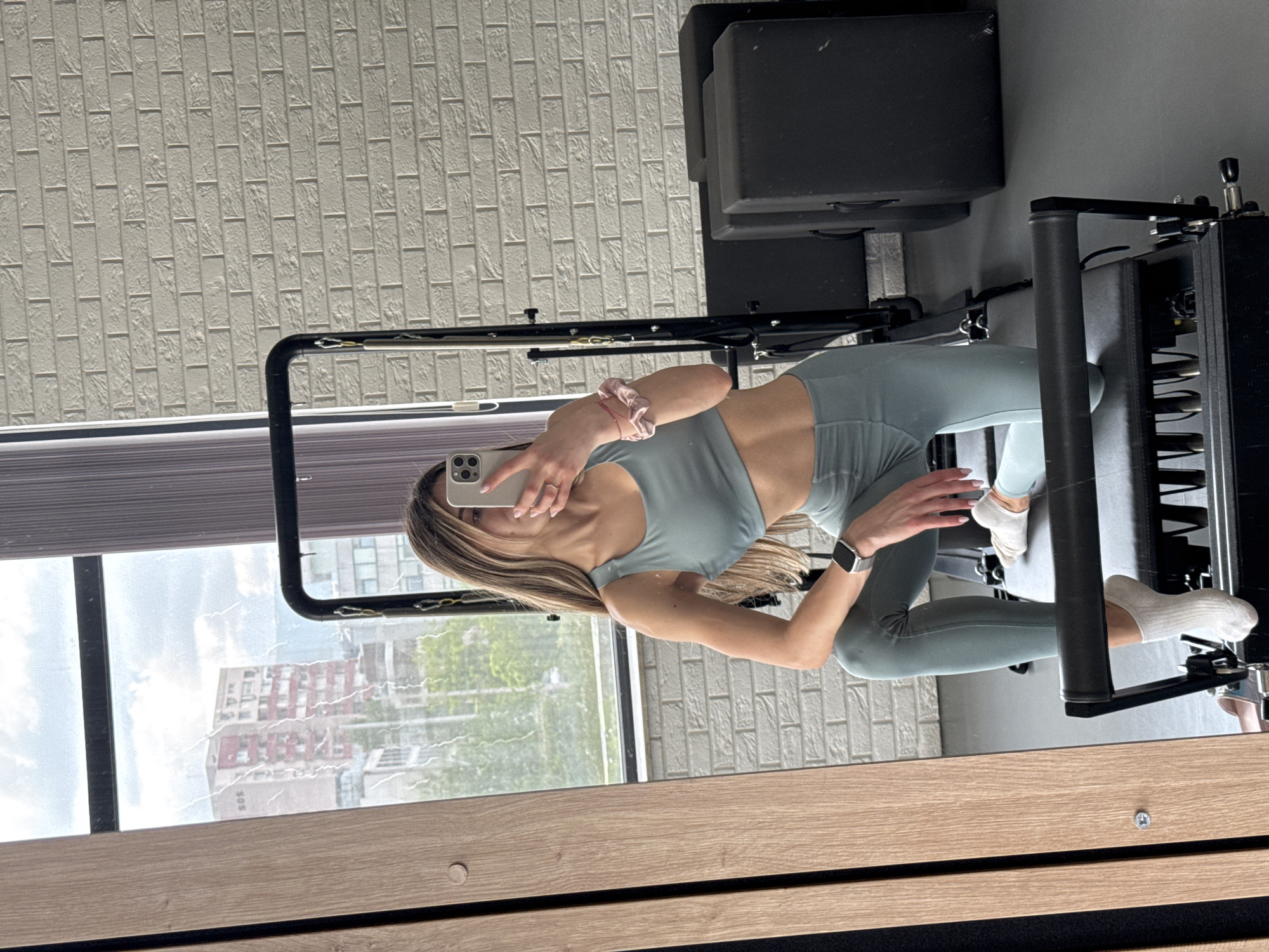 A fair-skinned woman with shoulder-length blonde hair is positioned in front of a pilates reformer, capturing a selfie using a smartphone. The woman is dressed in a matching athletic set with a sports bra and leggings, both in a muted sage green color. She is wearing white ankle socks and a silver smartwatch on her left wrist. A pink scrunchie adorns her left wrist, and a simple ring is visible on her right hand.
The pilates reformer is black and silver and partially obscures the woman. In the background, a large window reveals an urban cityscape. The buildings visible outside the window suggest the location may be Sofia, Bulgaria. Light is reflecting from the window, which could distort some details.
To the left of the woman, a wall is visible, textured with what appears to be a brick pattern. The bottom portion of the image shows a section of light wood paneling, possibly a desk or part of the reformer structure. A black block-shaped object sits behind the pilates reformer.
The woman is positioned such that her body is turned towards the reformer, while she holds her phone up to take the photo. The composition is a mix of indoor fitness setting with a view of the city.