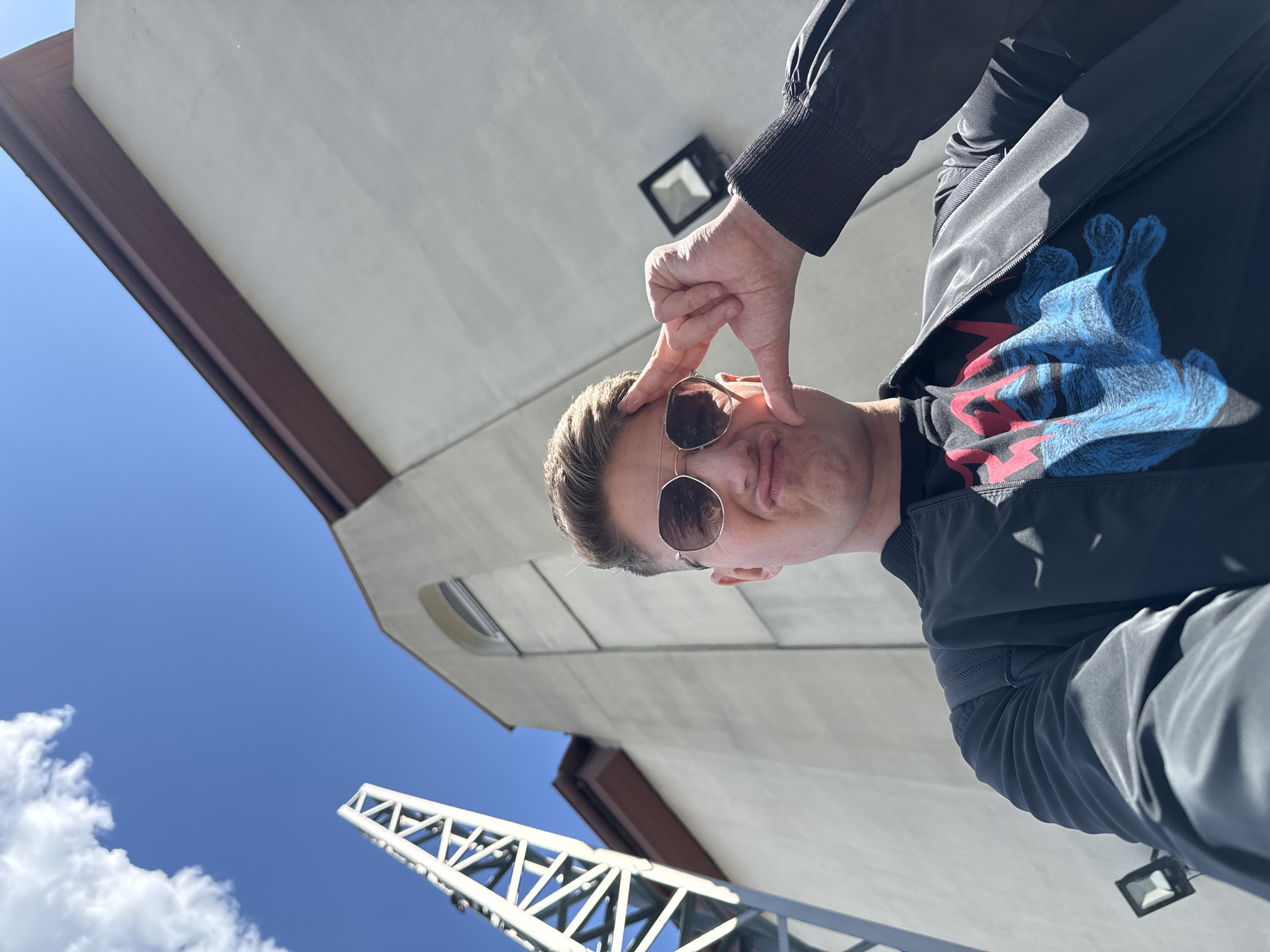 A man with blond hair and sunglasses makes a face for the camera, pinching his cheek. He wears a black t-shirt with a blue and red graphic design and a black jacket with a ribbed cuff. His left hand is visible framing his face. The background features a low-angle view of a light beige building facade with dark brown trim, a visible light fixture, and a steel structure against a blue sky with scattered white clouds. This location may be in Plovdiv, Bulgaria.