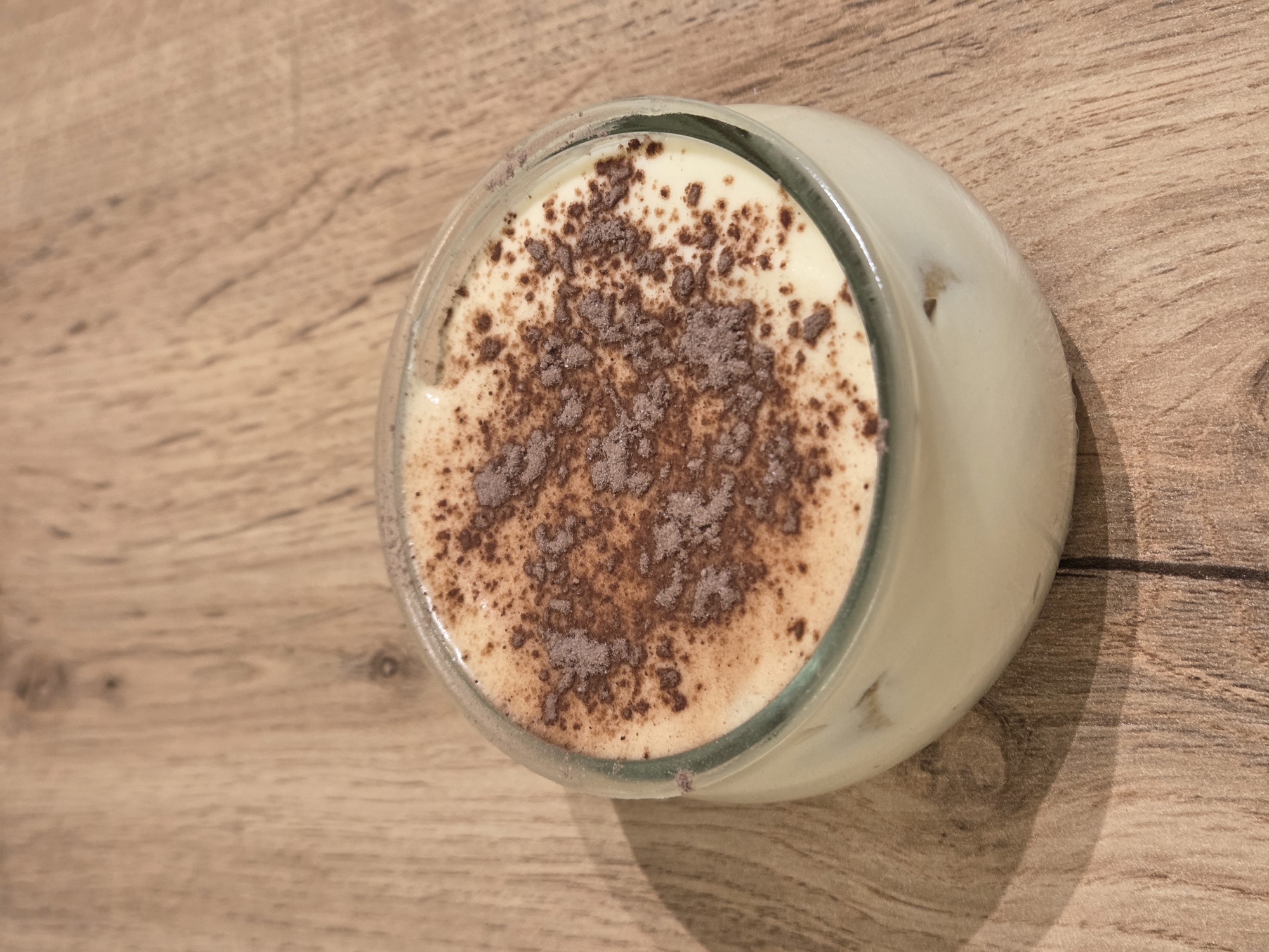 The image shows a top-down view of a dessert in a glass jar, sitting on a wooden surface. The dessert appears to be a light-colored cream or custard, topped with a dusting of dark brown powder, likely cocoa or chocolate. The glass jar is round with a slightly flared rim. The wooden surface has a light brown, woodgrain texture. The background is out of focus, maintaining the focus on the dessert. There are no people visible in the image. There are no specific clues to suggest that this is in Varna, Bulgaria.