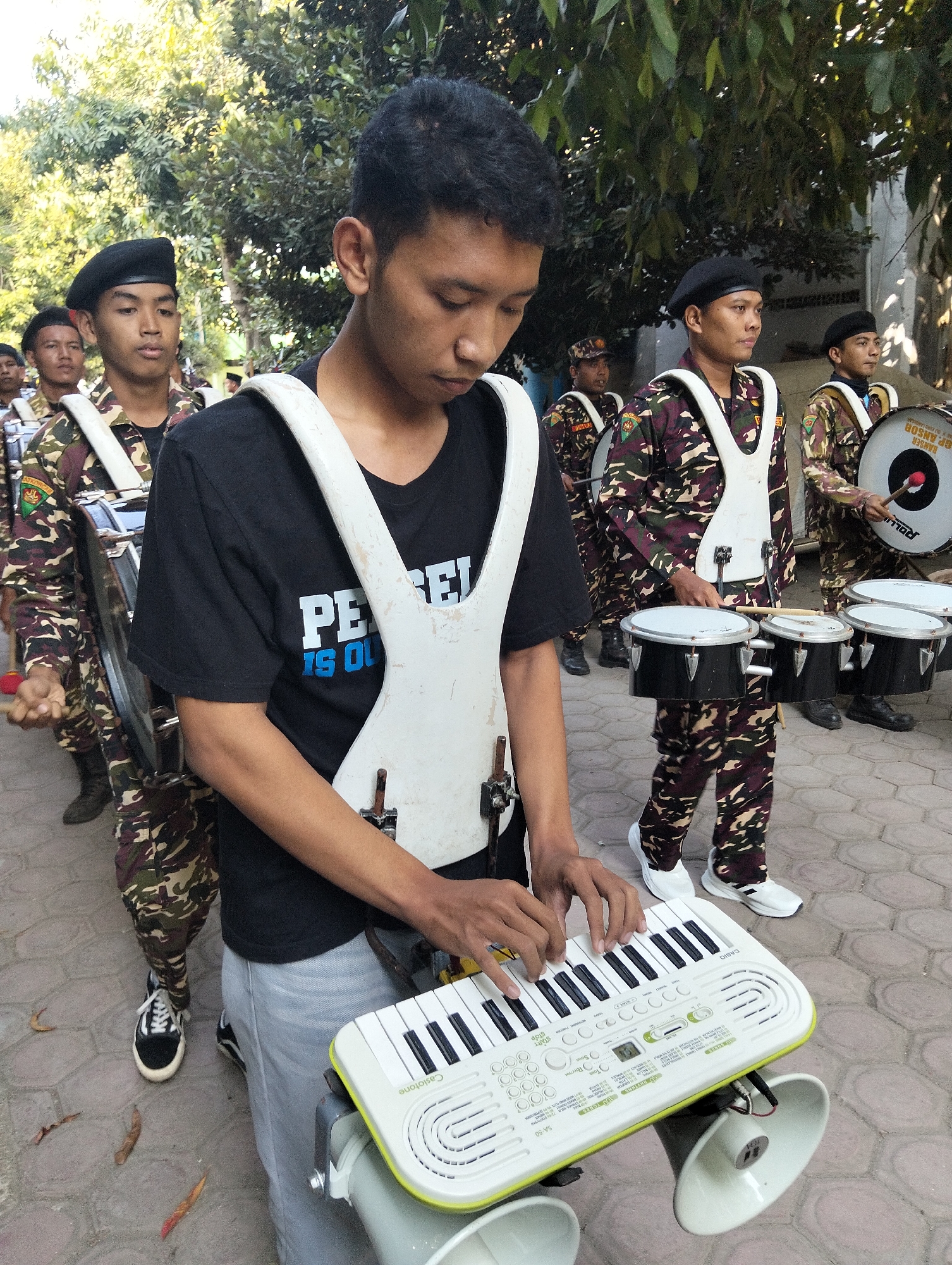 In Kranji, Indonesia, a group of young men are participating in a parade. The primary subject, in the foreground, is playing a small keyboard with speakers attached. He is wearing a black t-shirt with white lettering that appears to read "PERSEI IS O". He also has a white harness over his t-shirt. To his left and further into the background, other participants are marching. They are dressed in camouflage uniforms with black berets, and some are carrying large drums. One individual is seen holding drumsticks. The ground is paved with light-colored bricks. Trees with green foliage are visible in the background, suggesting an outdoor urban or suburban setting. The lighting indicates it is daytime.