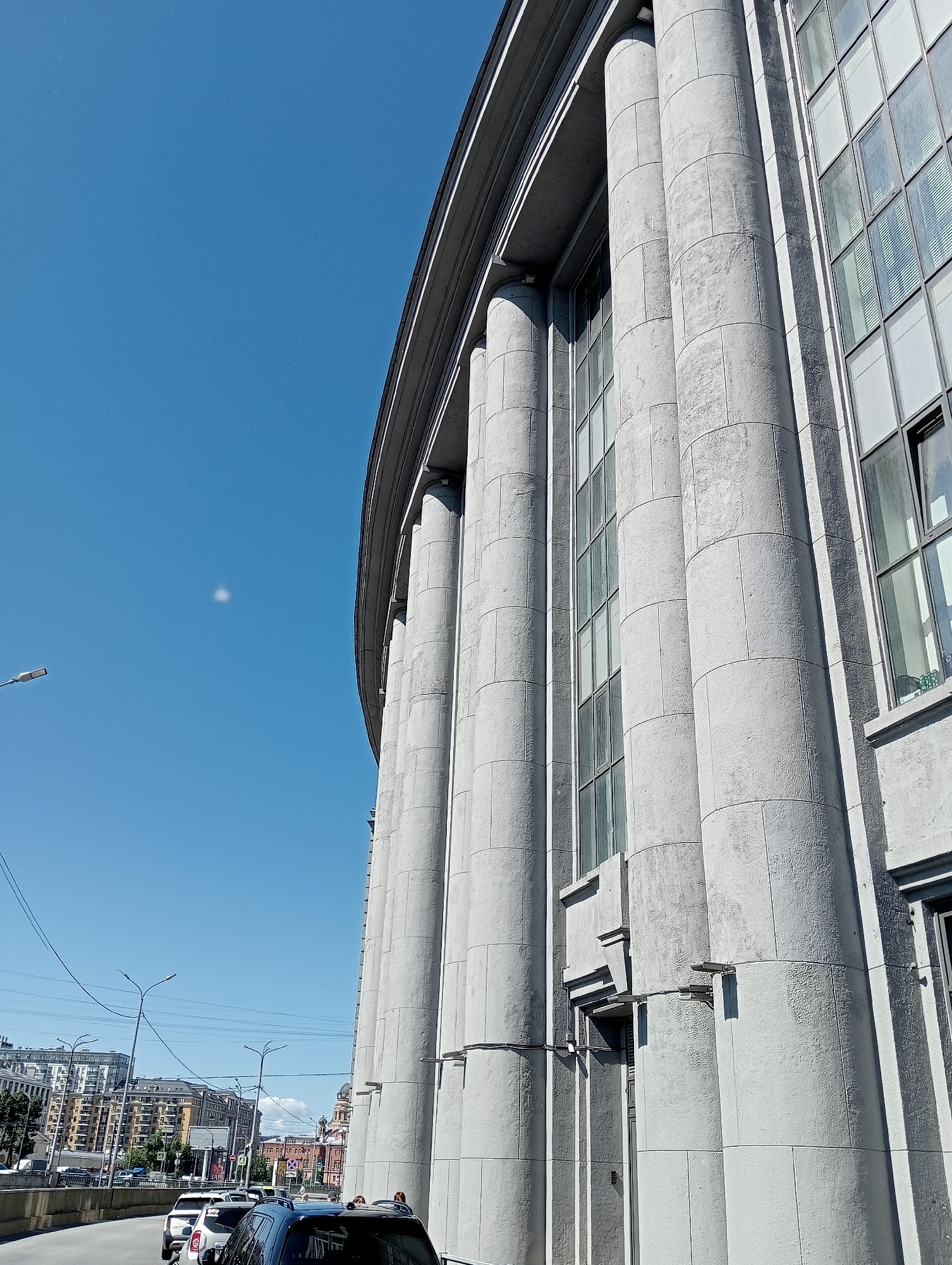 The image shows a view of the Singer House (Dom Knigi) in Saint Petersburg, Russia, seen from a low angle looking upwards towards the sky. The facade of the building is dominated by a row of massive, fluted columns made of light gray stone. The columns are cylindrical and appear to be constructed from segmented blocks. Between the columns are tall, multi-paned windows with blinds. The building's cornice runs along the top.
To the left of the Singer House, the sky is a clear, bright blue with a faint, diffuse white object, possibly a lens flare or a distant cloud. A street light with a horizontal fixture is visible on the left side of the frame, pointing upwards and to the left. In the background, a city skyline is visible with various buildings, including residential apartment blocks and older structures with domes. A road or embankment runs in the foreground, with several cars parked or in traffic. Some street lights and overhead power lines are also present. The overall impression is a sunny day in an urban environment.