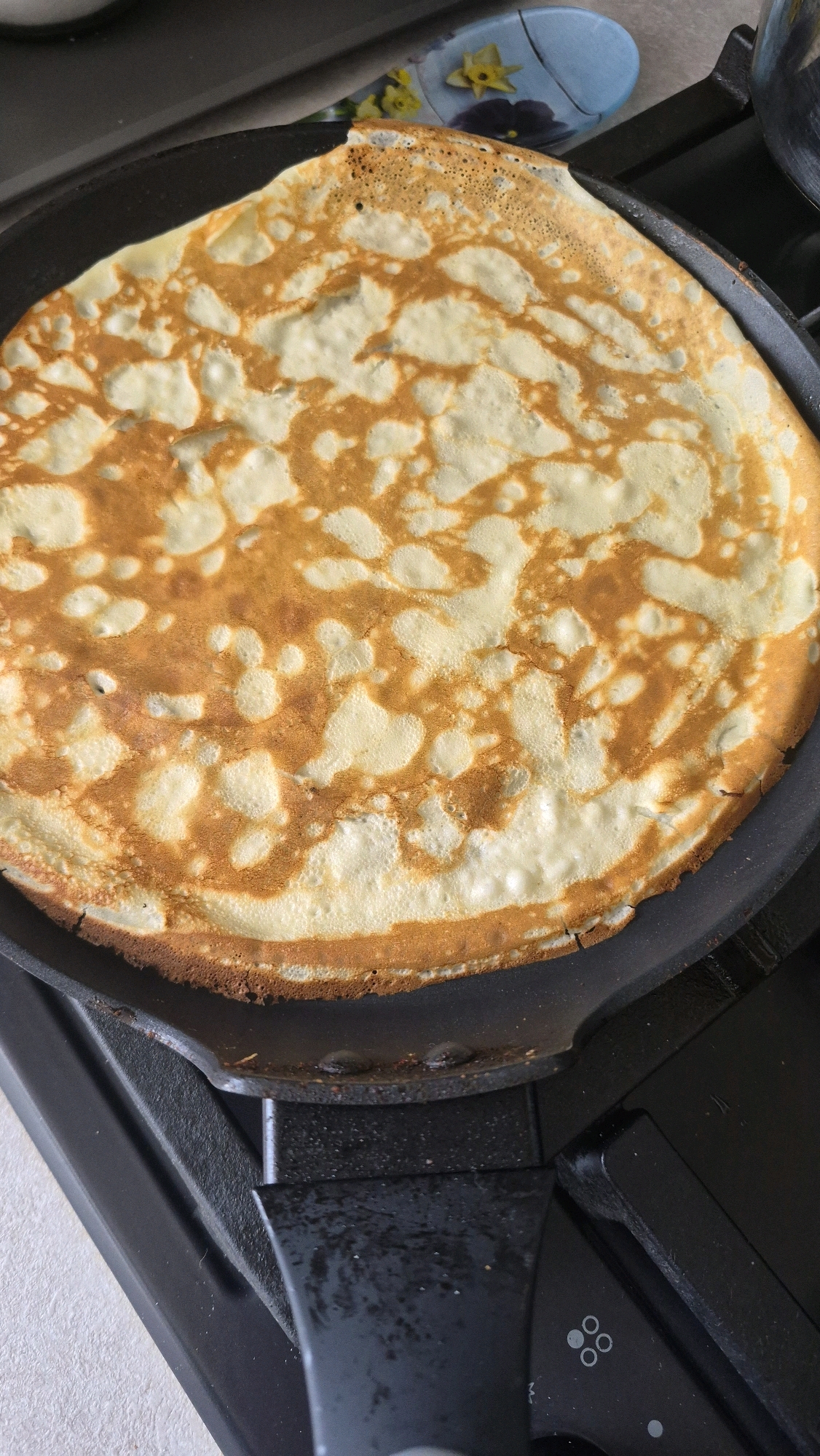 A golden-brown crepe is being cooked in a black non-stick pan on a black stovetop. The crepe has a lacy, irregular pattern of lighter and darker cooked areas. The edge of the crepe is slightly crisp and browned. A blue and yellow decorative object with floral patterns is visible in the background, to the upper right of the crepe. The stovetop has control knobs or indicators, one of which shows a circular pattern of dots. The scene appears to be an indoor kitchen environment, possibly in Krauja, Latvia.