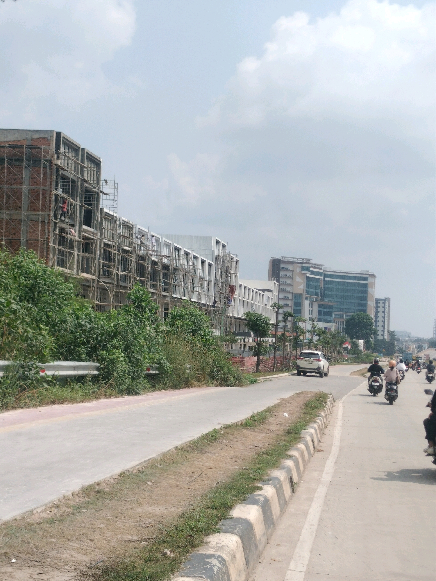 The image captures a street scene in Batam, Indonesia, likely in a developing area. On the left, a multi-story building is under construction, with scaffolding and workers visible. The building appears to be a modern commercial or residential complex, with exposed brickwork and concrete structures. In front of the construction site, a row of lush green trees and bushes lines the road.
The road itself is paved and appears to be a main thoroughfare. Several motorcycles are traveling on the right side of the road, indicating typical traffic flow in the region. A white car is also seen driving in the same direction. In the background, a sleek, modern high-rise building with glass facades stands prominently, suggesting a more developed part of the city. The sky is partly cloudy, with a bright, hazy quality typical of a tropical climate. The overall impression is one of urban development and ongoing construction in Batam.