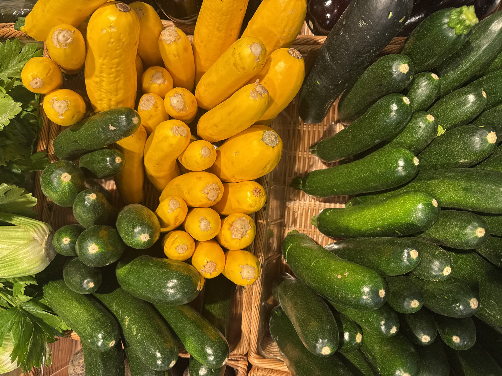 This is a close-up, overhead shot of a basket overflowing with a variety of fresh vegetables. The primary subjects are zucchini, yellow squash, and some celery stalks. The vegetables are arranged in a somewhat chaotic yet appealing manner, filling the woven basket.
The scene appears to be a market or grocery store display, with the vegetables neatly presented for sale. There are no people visible in the image, and the focus is entirely on the produce. The lighting suggests an indoor setting, possibly under artificial lights, as there are no strong shadows indicating natural daylight. The colors are vibrant, with the deep greens of the zucchini and celery contrasting with the bright yellow of the squash.
Notable details include the textured skins of the squash and zucchini, some showing slight imperfections or blemishes that are characteristic of fresh produce. The ends of the squash have a light brown, dried appearance. The celery stalks are a lighter green and have a crisp, leafy texture at the top. The basket itself is made of woven material, likely wicker or straw, which adds a rustic feel to the presentation.
There is no visible text in the image. The overall impression is one of abundance and freshness, showcasing a colorful selection of healthy ingredients. The location context of Höhenkirchen-Siegertsbrunn, Germany, suggests this could be a local farmer's market or a grocery store within that region.