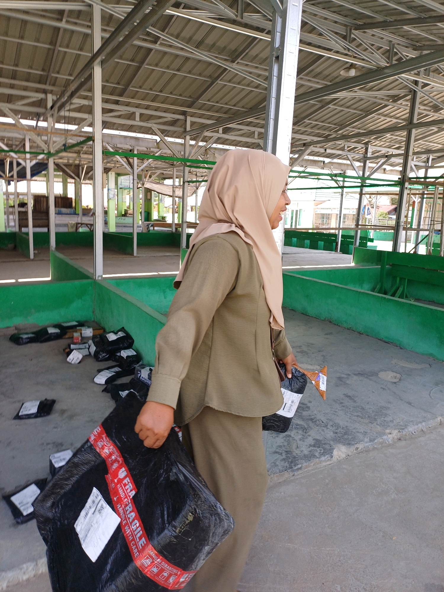 A woman wearing a beige hijab and a matching long-sleeved outfit stands in an open-air market structure in Pasir Belengkong, Indonesia. She is holding a large black plastic bag secured with red and white "FRAGILE HANDLE WITH CARE" tape in her right hand, and a smaller brown package in her left hand. The ground is concrete, and the market structure has a metal roof supported by metal beams. Several smaller black packages are scattered on the ground in the background. The market appears to be mostly empty, with green painted walls and benches visible in the background. The lighting suggests it is daytime.