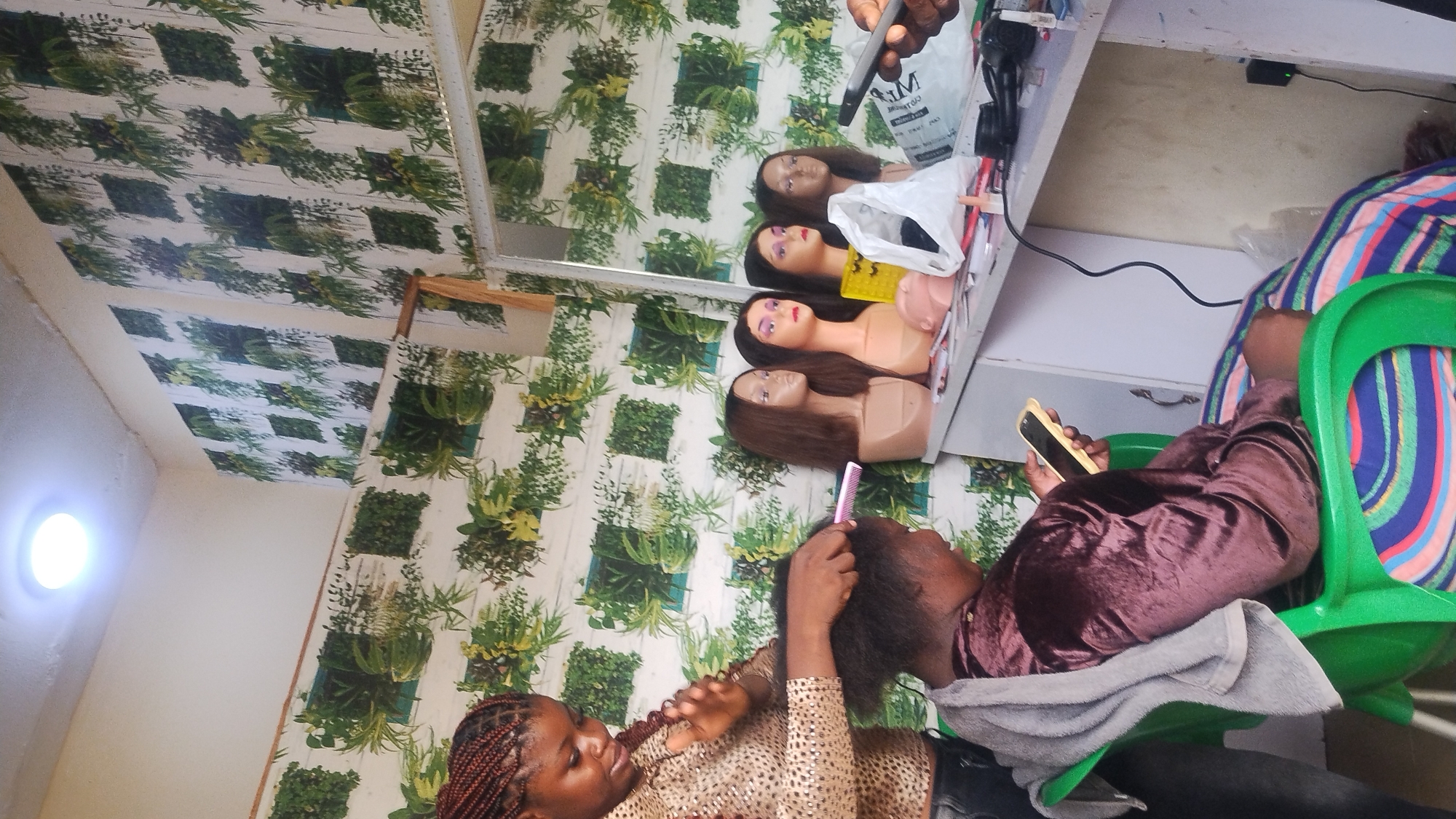 Inside a room in Jos, Nigeria, a woman with brown braided hair, wearing a brown and gold patterned shirt, stands and combs the hair of a seated person. The seated individual, wearing a brown satin outfit, reclines in a green plastic chair with a colorful striped cushion, holding a gold-colored smartphone. A gray towel is draped over the chair. Behind the seated person, a white desk holds three mannequin heads with wigs, and a white shopping bag is visible. A mirror hangs above the desk, reflecting the mannequins and the wall, which is covered in a green plant print wallpaper. A bright ceiling light is visible in the corner of the room.