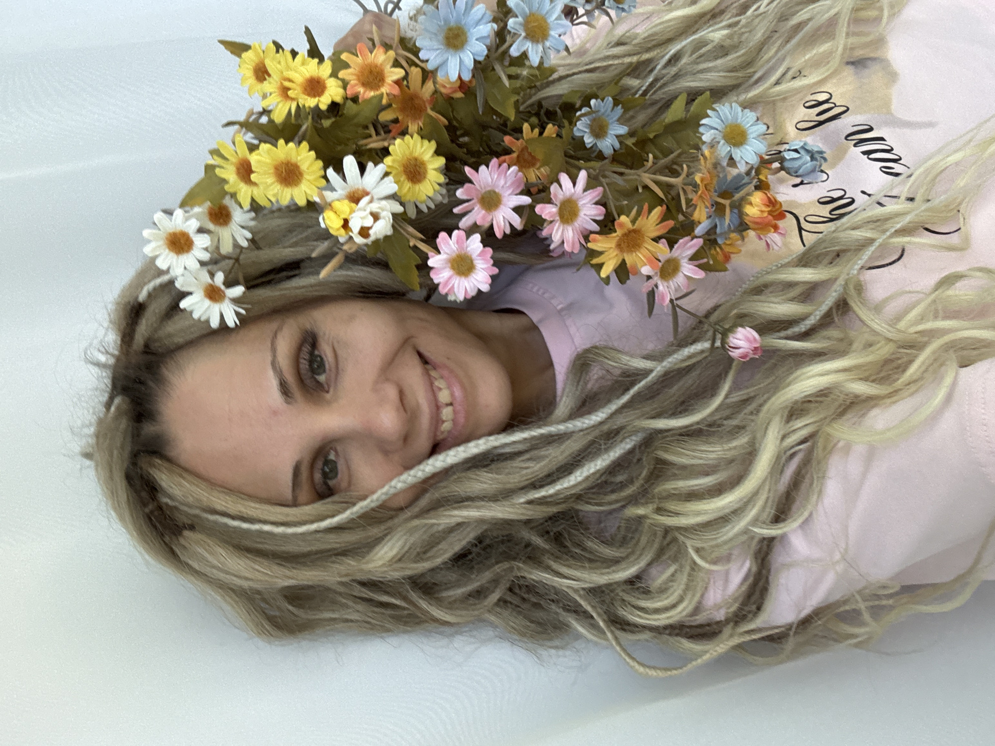 A woman with long, wavy blonde hair with a few white braids lies on her back on a white surface, with a bouquet of colorful daisies (white, yellow, pink, blue, and orange) resting on her head and partly covering her face. She is wearing a light pink top. She is smiling and looking at the camera. The location is not discernible from the image.
