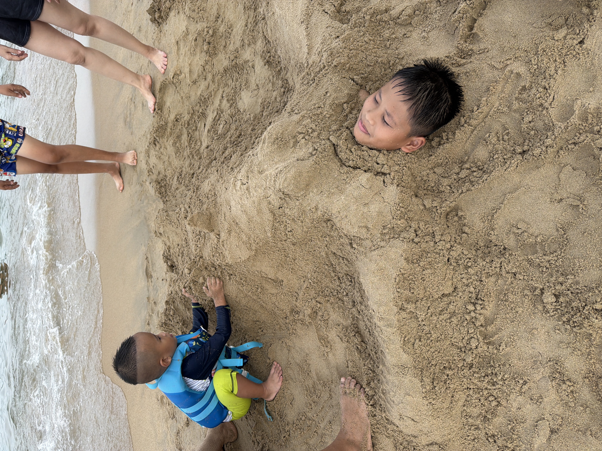 A young boy wearing a life vest and swim trunks is playing in the sand near the ocean. His head is buried in the sand, with only his face exposed. Another child is partially visible in the background, standing by the water's edge. The scene takes place on a sandy beach, likely in Nha Trang Ward, Vietnam, given the context of the question. The water is calm, with gentle waves lapping at the shore. The sun appears to be shining, casting a warm glow on the scene.