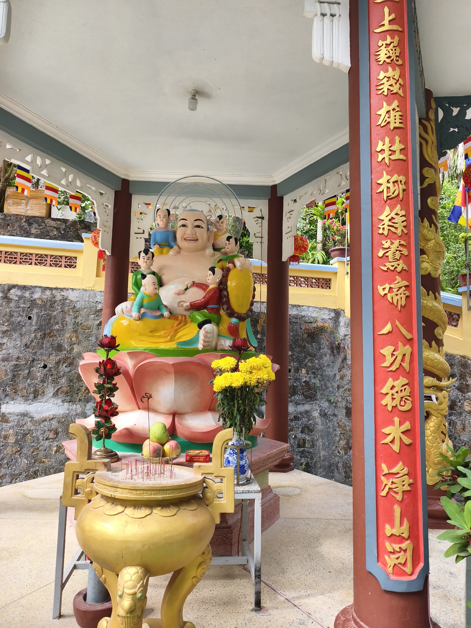 The image depicts a colorful Buddhist shrine located in the city of Tay Nha Trang Ward, Vietnam. At the center is a large, smiling Buddha statue surrounded by children. The statue sits atop a lotus flower base. In front of the statue is a golden incense burner filled with incense sticks, accompanied by offerings of fruit and flowers. A red and gold-ornamented pillar with Chinese script stands to the right. The shrine is situated within a covered structure, with decorative elements and flags visible in the background. The location appears to be a hillside temple or religious site.