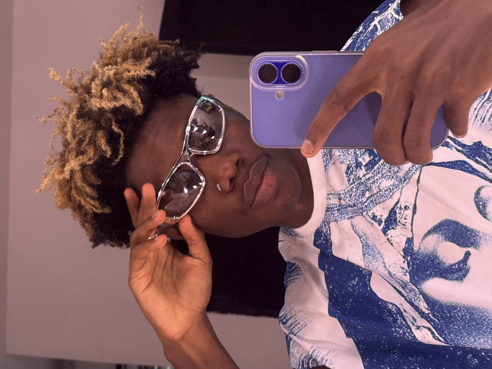 A person with dark skin, blond-tipped hair, and silver sunglasses is taking a selfie with a lavender-colored smartphone. They are wearing a white shirt with a blue abstract pattern. The background appears to be a room with a plain wall and dark fabric hanging. The location is identified as Igboefon, Nigeria.