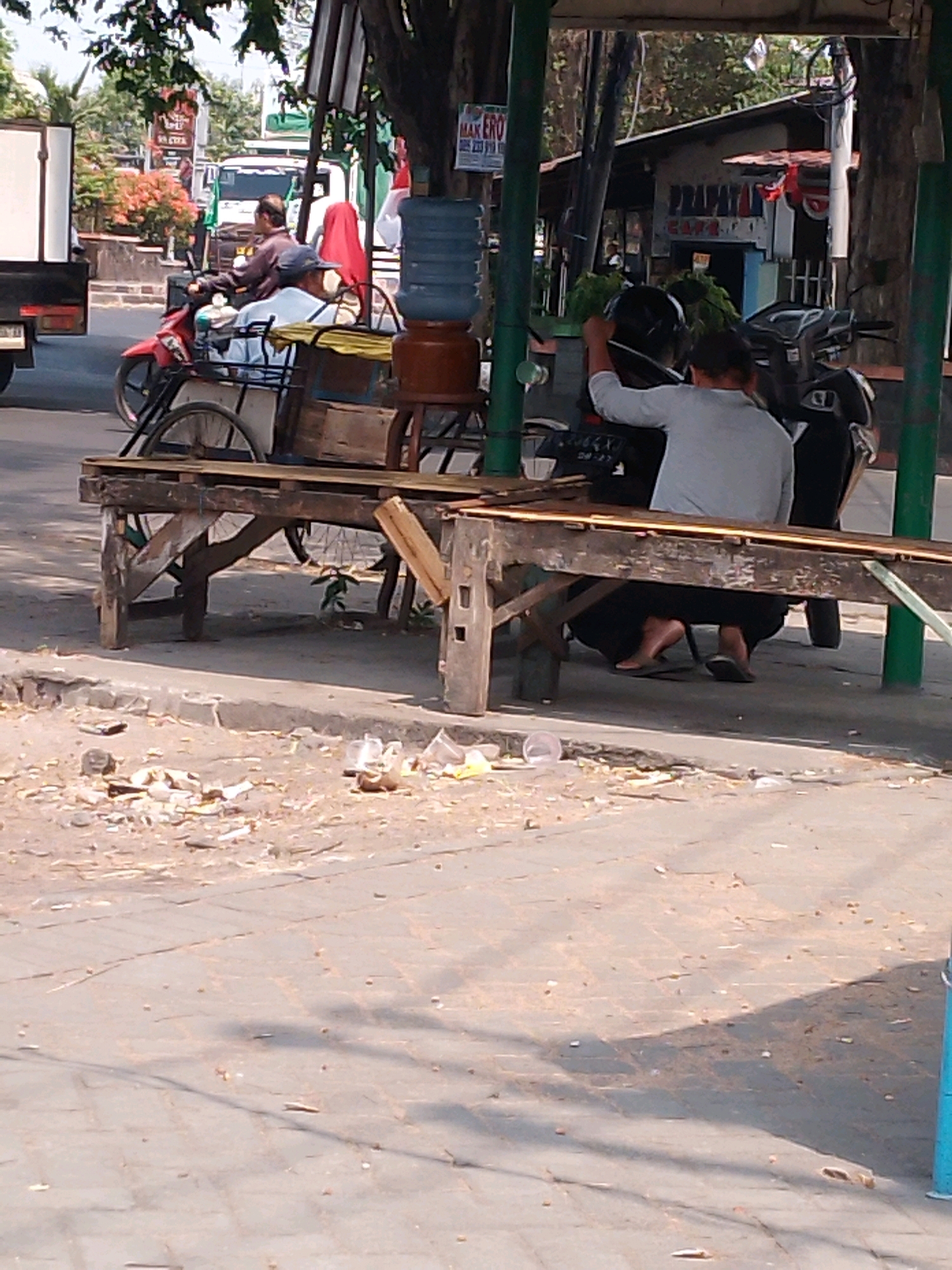 This image captures a street scene in Kutorejo, Indonesia, under a canopy of trees. In the foreground, a weathered wooden bench or stall sits on a cracked, dusty pavement littered with debris, including plastic bottles and wrappers. Behind the bench, a man wearing a light grey long-sleeved shirt and dark pants is seated, interacting with a motorcycle parked beside him. He appears to be holding a dark helmet. Another motorcycle is parked to the left of the man.
In the background, a red motorcycle with a passenger wearing a red headscarf and a person in a blue shirt is visible, suggesting ongoing traffic and activity. Further back, a white truck and various buildings are present, indicating an urban or semi-urban environment. The overall impression is of a bustling roadside scene with everyday activities taking place.