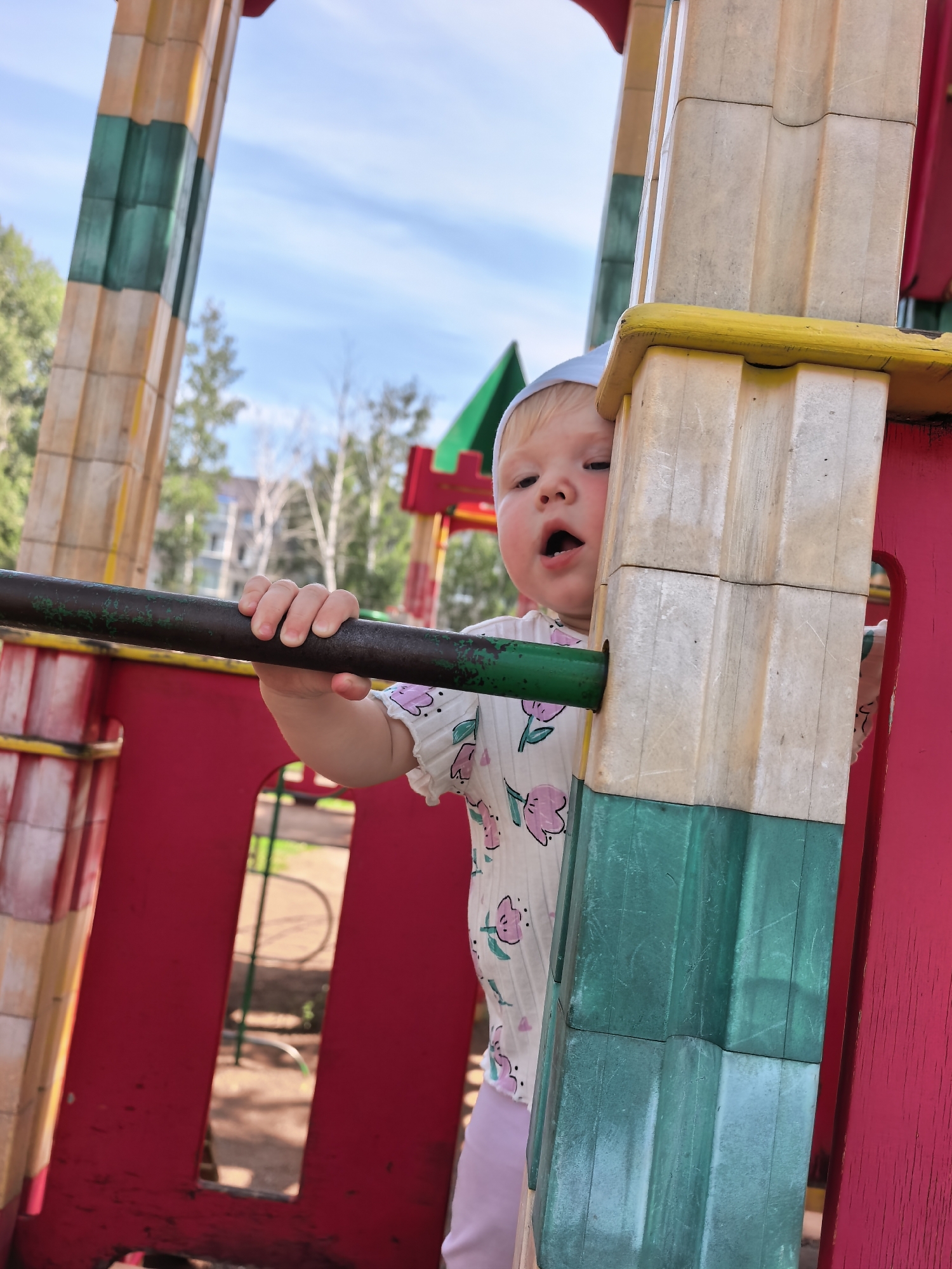 A Caucasian toddler, wearing a light blue cap and a white t-shirt adorned with pink floral patterns, peeks around a beige and green playground structure. The toddler's mouth is open, and their left hand is grasping a dark green metal bar. The background reveals a bright blue sky with scattered clouds, and the presence of trees and buildings suggests an outdoor urban setting. The playground equipment is predominantly red and features wooden-patterned vertical supports. The image appears to have been taken on a sunny day. The child's age is approximately one to two years old. The location of this playground is in the city of Sterlitamak, Russia.