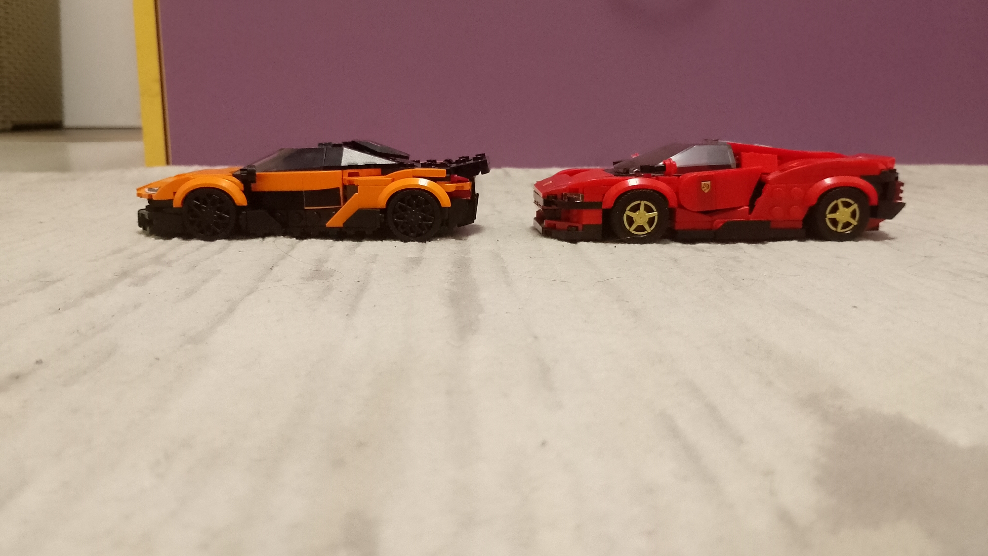 Two toy cars, likely brick-built models such as Lego Speed Champions, are prominently displayed on a light-colored, textured surface, possibly a carpet or bedspread. The cars are positioned facing each other at a slight angle, appearing as if in a static display.
The car on the left is predominantly orange with black accents on its lower body, roof, and rear spoiler. It features black wheels and has a sleek, aerodynamic design reminiscent of a modern supercar like a McLaren.
The car on the right is bright red with black elements along its lower edge and chassis. It sports distinctive gold-colored wheels and a small, golden emblem on its side, which closely resembles the iconic Ferrari logo. This car also exhibits a supercar-like silhouette.
The setting is an indoor environment. Behind the cars, a solid purple wall serves as the primary background. To the far left, a portion of a lighter, possibly white or off-white, wall is visible, along with a vertical yellow object, which could be a book spine or part of a furniture item. The lighting suggests an artificial indoor light source. There are no people visible, and no specific time of day or weather conditions are discernible. The scene presents a collection or playful arrangement of toy vehicles within an indoor space in Blagoevgrad, Bulgaria.