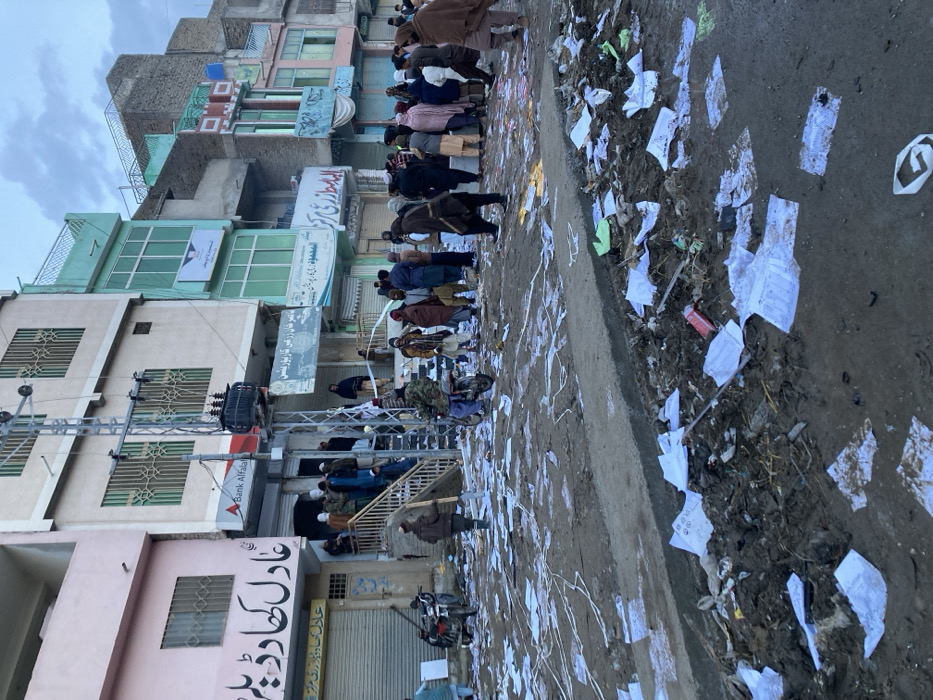 A gathering of people appears to be praying in a street lined with buildings. Debris and scattered paper cover the street, giving it a messy appearance. The buildings along the street are varied in color and show signs of wear. A sign for "Bank Alfalah" is visible, along with other signs in a different script. A motorcycle is parked near the bottom of the frame. The image has an overall feeling of urban grit and activity.