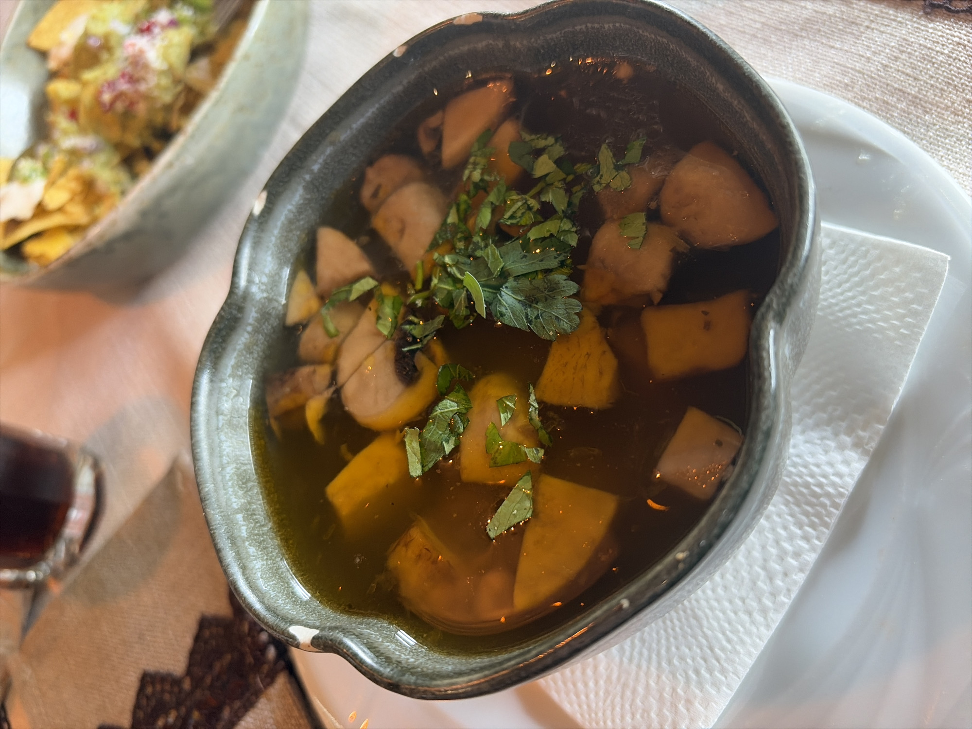 A high-angle, medium shot shows a bowl of food with chopped potatoes and herbs in broth on a white plate. The bowl is gray-blue with a dark rim. A white paper napkin sits between the bowl and the plate. To the left of the bowl, a partially visible bowl containing chips and guacamole is present. Further to the left, a glass of dark liquid sits on a patterned tablecloth. The food is possibly at a restaurant in Veliko Tarnovo, Bulgaria.
