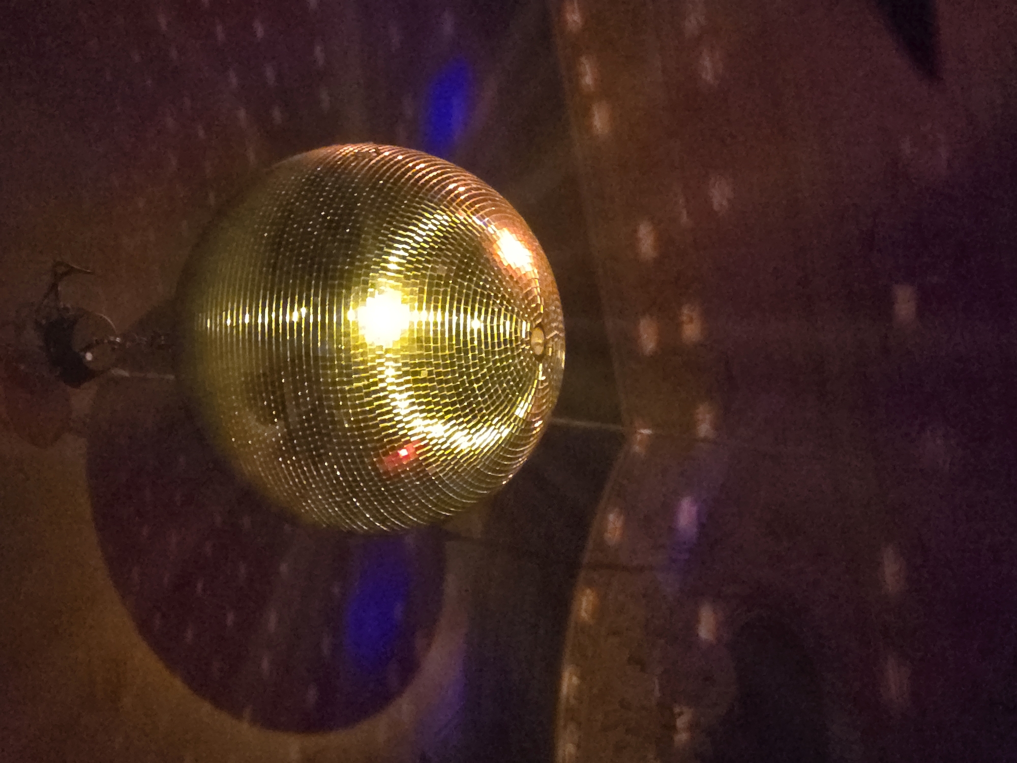 A disco ball is mounted on a wall in a dimly lit space. The ball reflects light onto the surrounding wall, creating spots of light. The wall appears to be textured. The location is not discernible from the image.