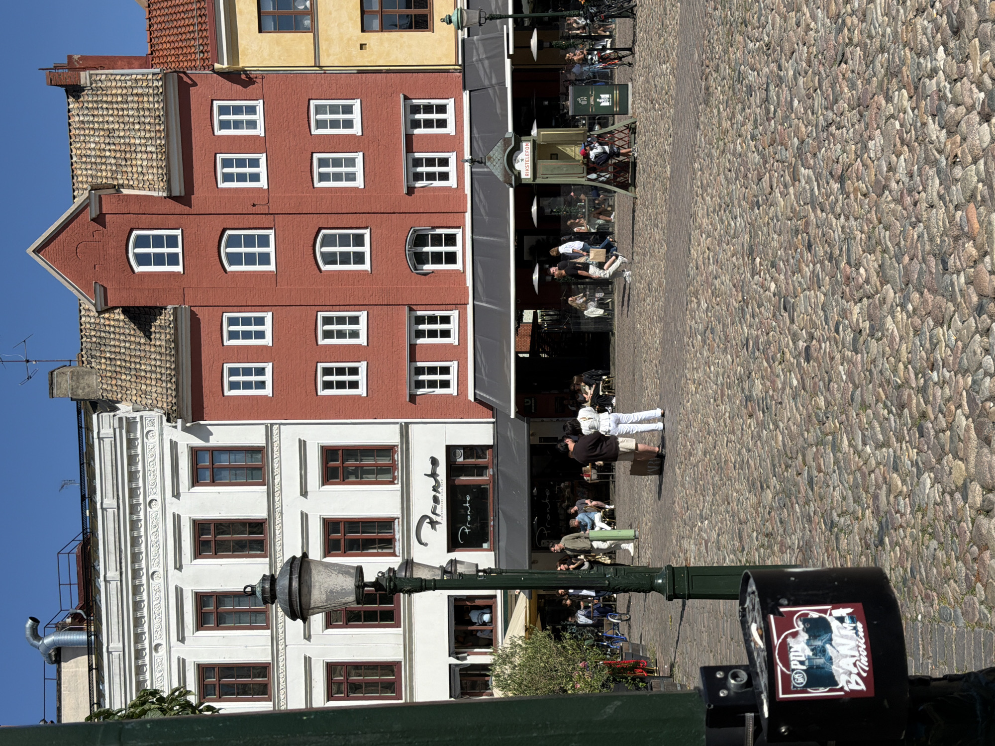 The image depicts a sunny day in a cobblestone plaza in Malmö, Sweden. In the foreground, a black trash receptacle with a sticker on it is visible, attached to a lamppost. Behind the receptacle, a streetlamp stands on a cobblestone surface. Several people are scattered throughout the plaza.
The background features a row of buildings. The leftmost building is white with decorative molding and multiple windows. Next to it is a red brick building with white-trimmed windows. To the right of that is a yellow building. A black awning extends across the lower level of the red and yellow buildings, covering outdoor seating areas for a restaurant or cafe named "Pronto." Several tables with umbrellas are set up, and patrons are seated.
A small, green structure with the words "RIKSSTELEFON" is positioned in front of the awning, likely a public telephone booth. In the middle ground, two individuals are standing near each other, one holding a suitcase. Other individuals are walking or sitting at the outdoor tables. Bicycles are visible parked near some of the buildings. The sky is a clear blue.