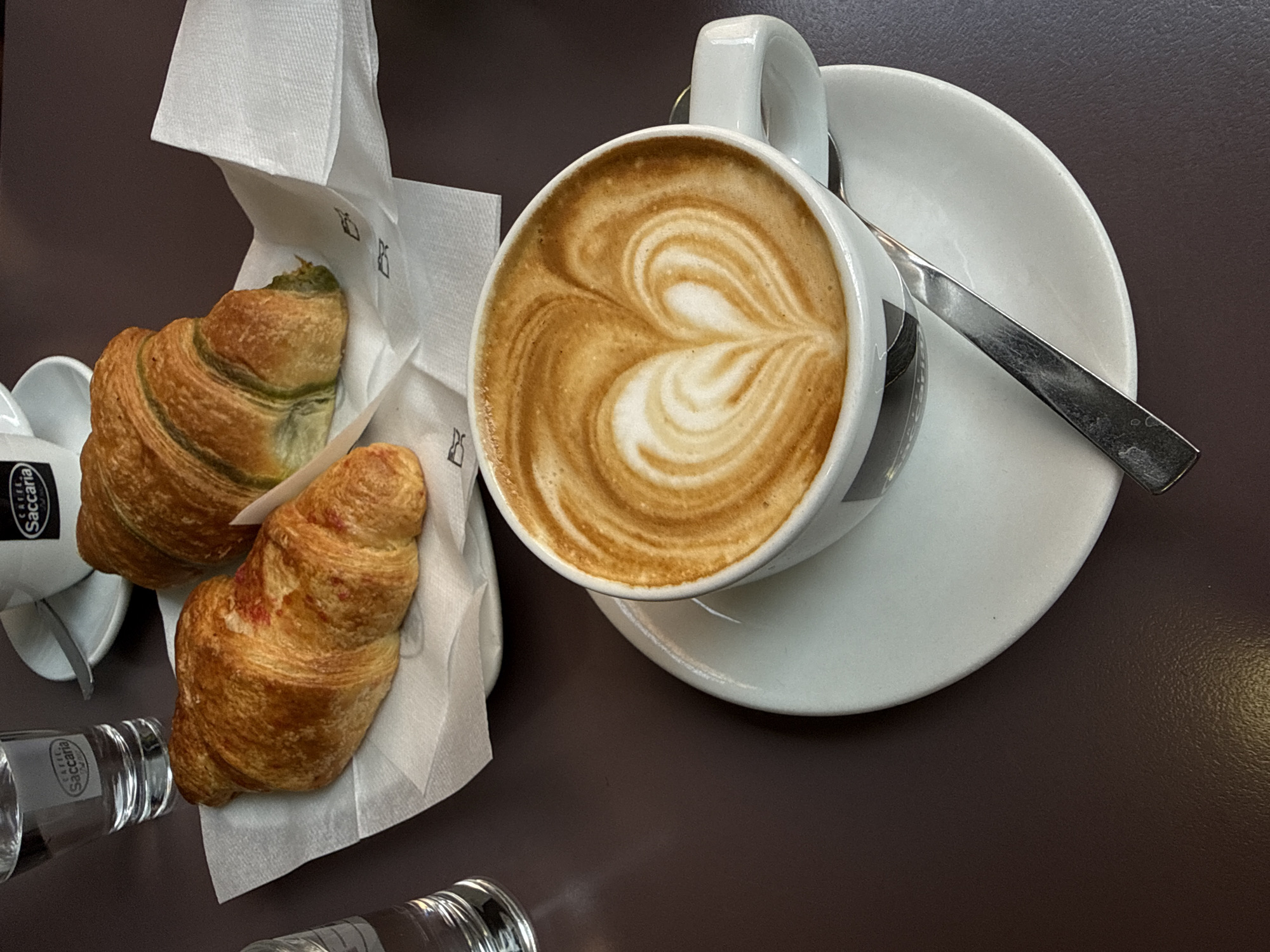 An overhead shot captures a delightful breakfast scene on a dark brown table, likely in Bologna, Italy, as suggested by the "Caffè Saccaria" branding on the cups. Two golden-brown croissants rest on a white paper napkin, one with a reddish filling visible, the other appearing to have a green filling. Beside them, a white cup and saucer hold a latte with intricate latte art, a heart shape formed by the milk foam. A silver spoon lies on the saucer, ready for use. Two glasses of water are partially visible on the left side of the frame. The lighting suggests an indoor setting, possibly a café, with soft shadows cast by the objects.