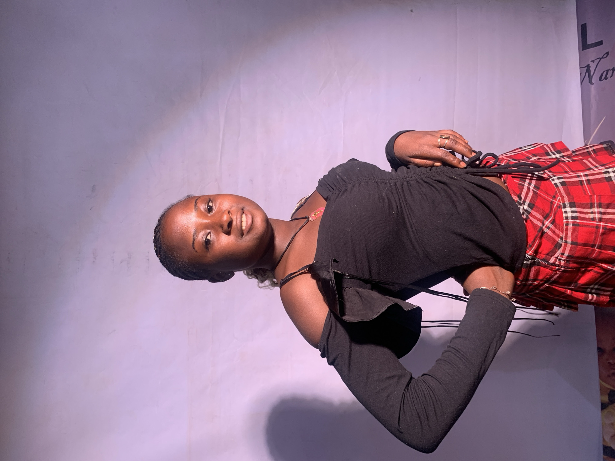 The image depicts a young woman against a backdrop, likely in a studio setting. She wears a black top with long sleeves and a red plaid skirt. Her hair is neatly styled. She is holding the drawstrings of her top. The environment is a plain backdrop. The location is possibly Jos, Nigeria but the photograph does not contain information about Jos or whether the photograph was taken in Jos.