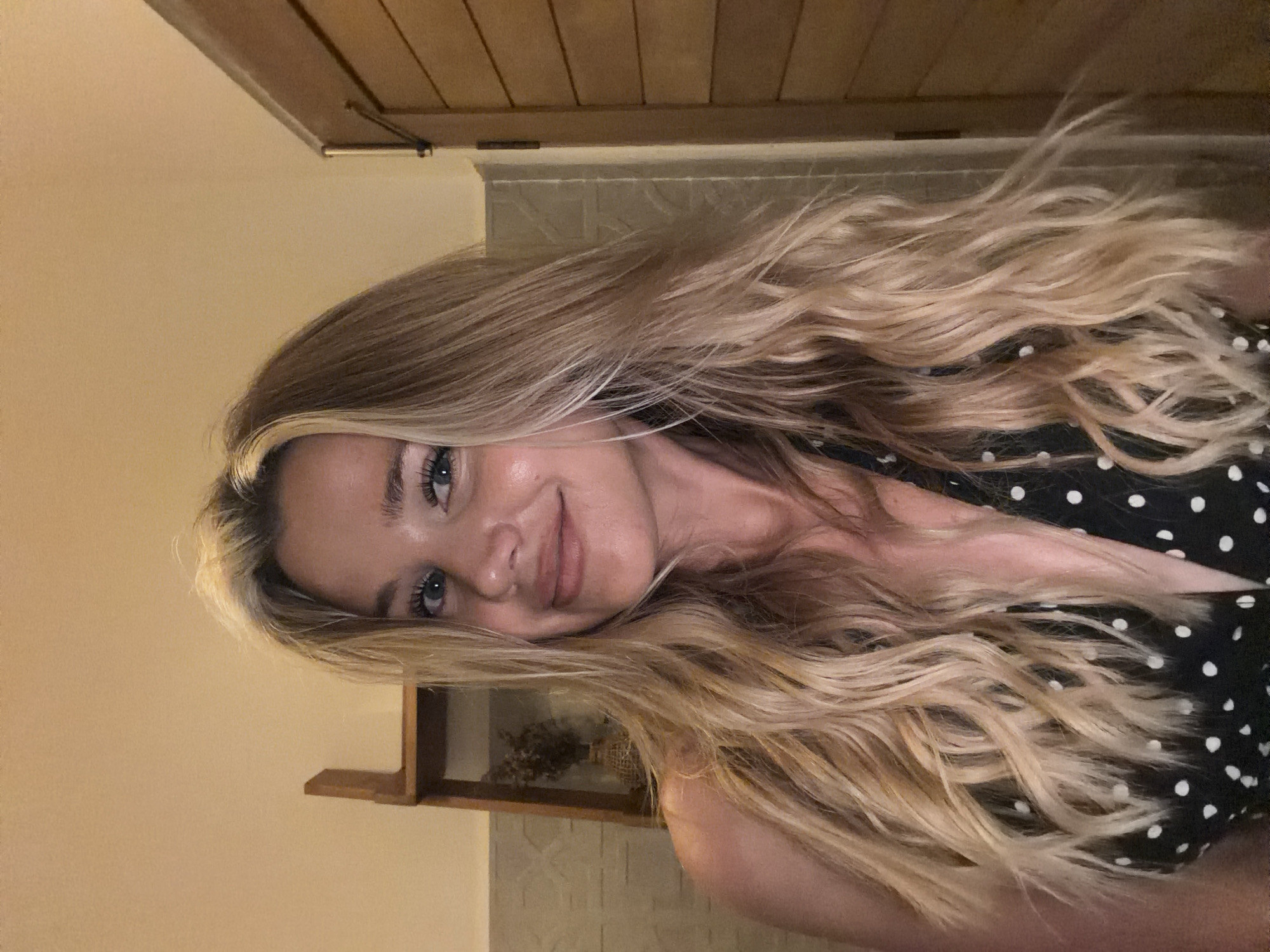 A medium shot shows a fair-skinned woman with long blonde wavy hair and blue eyes, smiling at the camera. She is wearing a black top with white polka dots. The background appears to be an indoor setting, possibly a bathroom or a room with wooden paneling on the right side. A wooden shelf with a vase containing dried flowers is visible on the left side. The lighting is warm and dim. The image appears to be taken in Choeng Thale, Thailand.