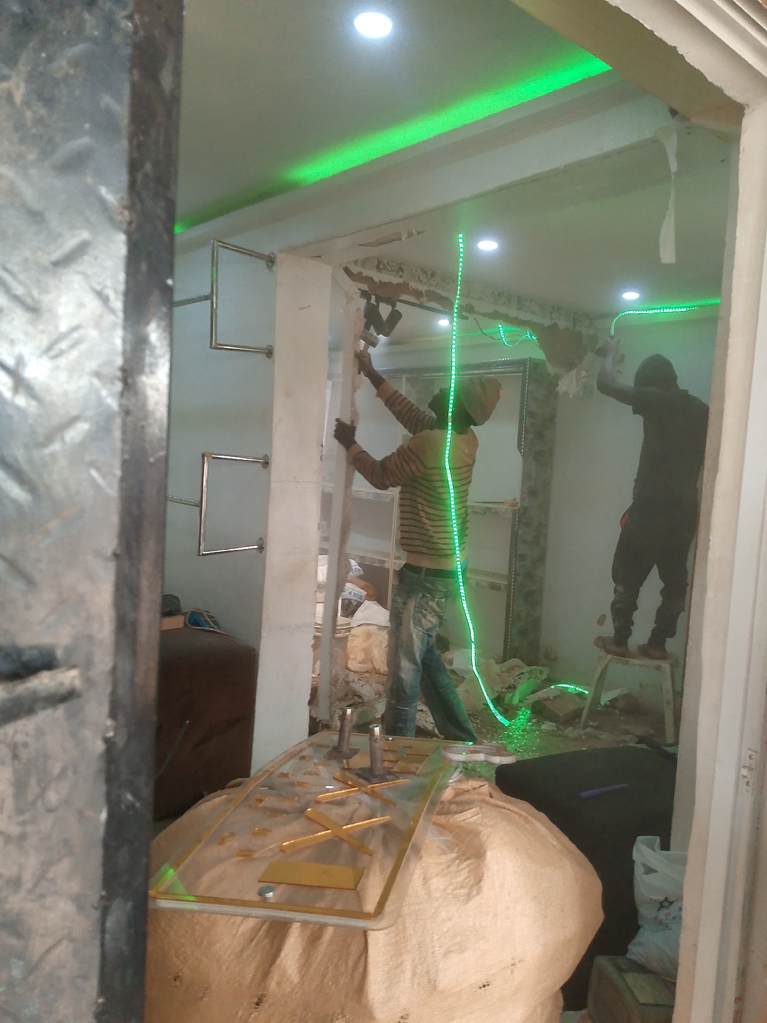 In the city of Bukuru, Nigeria, two individuals are engaged in construction work within an interior space that appears to be undergoing renovation. The room is illuminated by overhead lights and prominent green LED strip lighting installed along the ceiling and a vertical plane.
One worker, wearing a striped long-sleeved shirt and jeans, stands to the left. He is positioned near a wall and appears to be holding or operating a tool, possibly for measurement or installation, as indicated by the vertical green laser line extending from his position.
The second worker is on the right, standing on a step stool. He is dressed in dark clothing and is also facing the wall, seemingly involved in the ongoing construction or decoration activities. His posture suggests he is reaching or working at an elevated level.
The environment shows signs of construction, with rubble and debris on the floor. There are also various construction materials and items present, including large sacks, possibly for carrying materials or waste, and a transparent sheet with golden metallic accents laid on top of one of the sacks in the foreground. Shelving units are visible on the left wall, and some metal fixtures are also present. The scene captures a moment of active renovation or building work in a confined indoor setting.