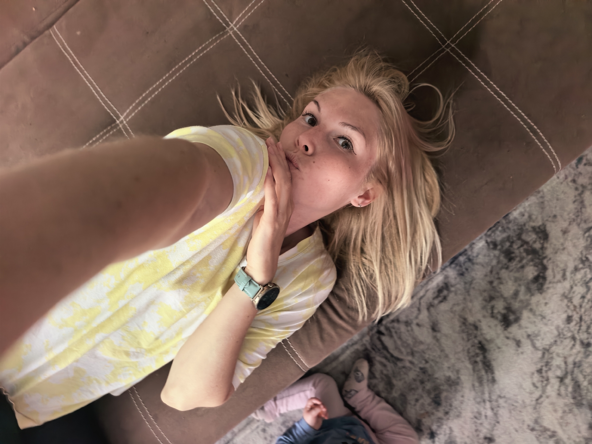 The image captures a high-angle, overhead perspective of a blonde woman lying on a brown, stitched couch. She appears to be taking a selfie, holding her arm extended towards the camera. The woman is wearing a yellow and white patterned t-shirt and a light teal smartwatch with a gold bezel on her left wrist. Her hand is positioned near her mouth, creating a puckered expression, possibly blowing a kiss, and she looks directly into the camera with a playful gaze. Her blonde hair is spread out around her head on the couch.
In the bottom right corner of the frame, a small child is partially visible, lying on a patterned grey and white rug next to the couch. The child is wearing pink pants and a blue top. The scene appears to be indoors, likely in a living area or bedroom, suggesting a moment of relaxation or play. The lighting indicates it is daytime. There is no visible text in the image.