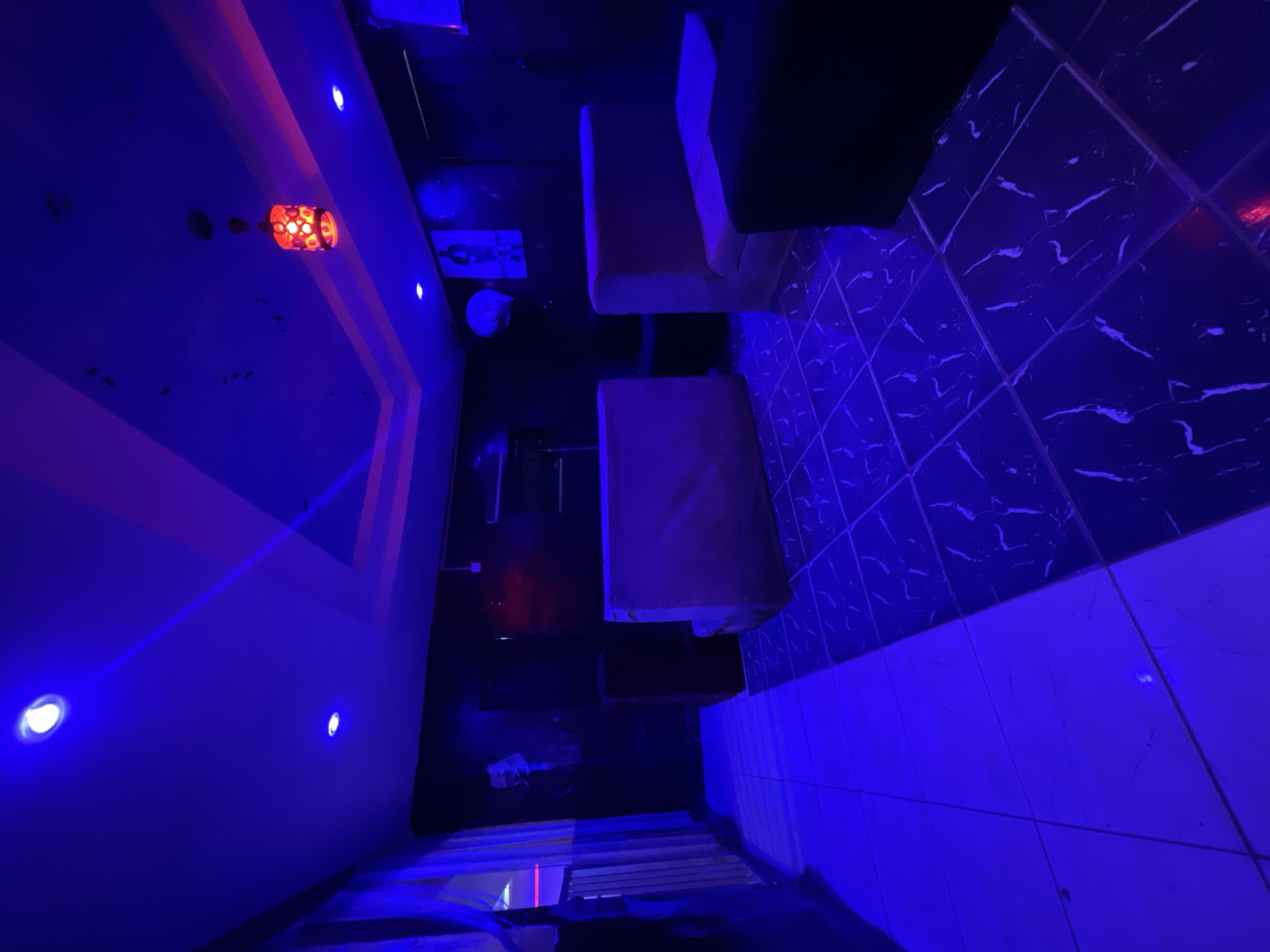 The image depicts a dimly lit lounge area, likely a cafe or bar, in Durumi, Nigeria. The space is illuminated by a combination of blue and pink lighting, creating a vibrant and moody atmosphere.
The room features dark, textured walls with some artwork visible. The floor is tiled in a black and white marble pattern, reflecting the ambient light. Several pieces of furniture are present, including what appear to be low-lying sofas or banquettes in neutral tones, and a dark-colored stool or ottoman.
A decorative pendant lamp with a red glow hangs from the ceiling, adding a contrasting warm element to the cool blue lighting. Recessed ceiling lights also contribute to the illumination. There's a large window or glass partition on the left side of the room, draped with sheer white fabric, through which hints of exterior light and color can be seen.
The overall impression is of a modern, stylish, and intimate space designed for relaxation and socializing. There are no people or any discernible actions taking place in the image.