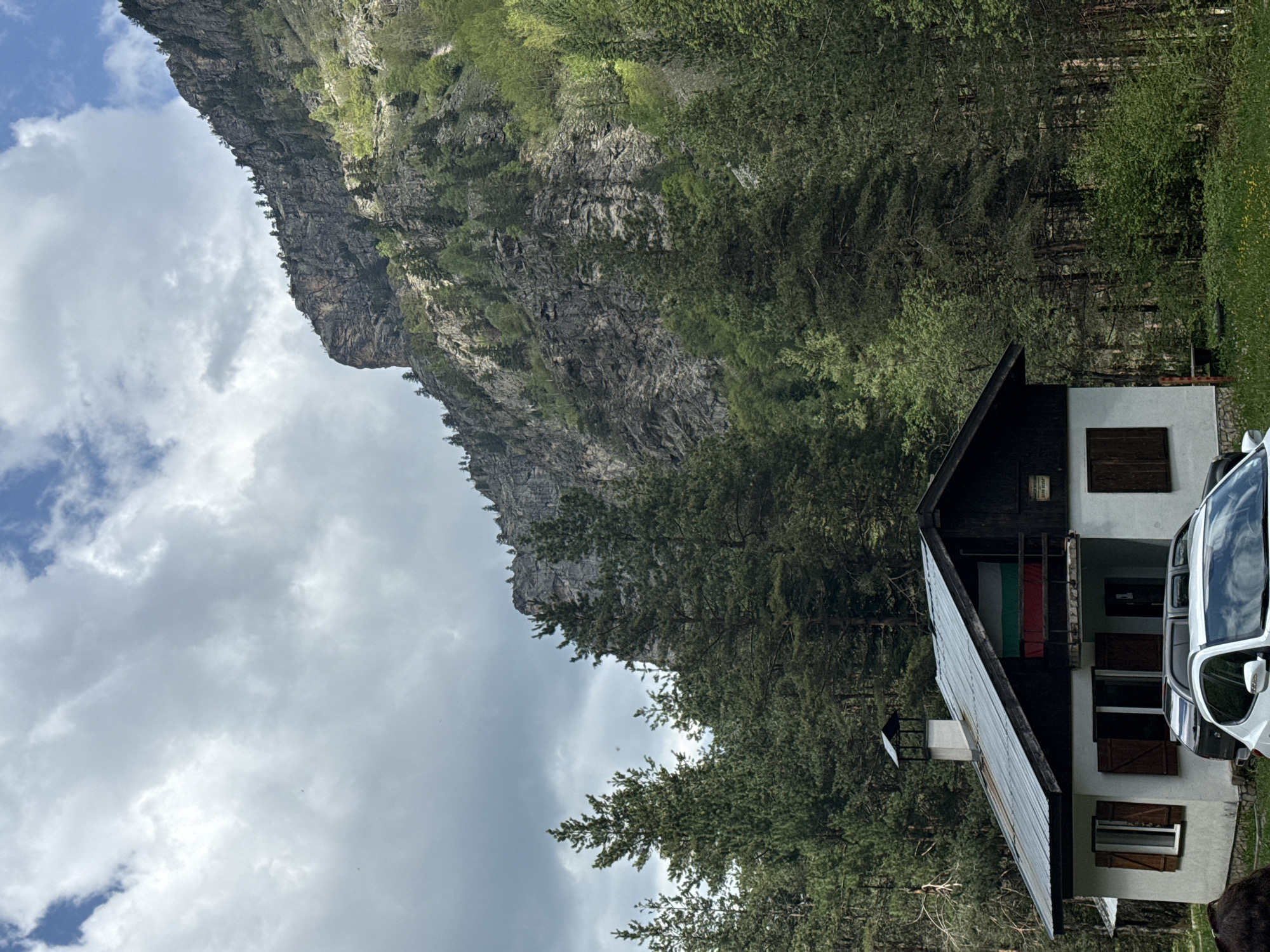 The image depicts a mountainous landscape in Beli Iskar, Bulgaria. A two-story house with dark wood accents and a metal roof sits amid tall pine trees. A white car is parked next to the house, and the Bulgarian flag is displayed on the house. Above the house is a tall cliff face with some trees growing on it. The sky is cloudy.