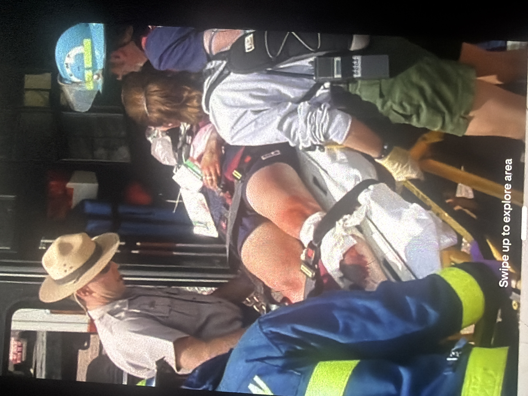 An image depicts a patient on a stretcher being attended to by several individuals. The patient, whose legs are visible, appears to be in distress, with some visible injuries. Two individuals in the foreground are wearing blue jackets with yellow reflective stripes, suggesting they are emergency responders. One individual in the middle ground wears a light-colored shirt and a straw hat, and another person in the background wears a blue hard hat. The scene appears to be outdoors, possibly near a vehicle, as indicated by the reflective surfaces and the general arrangement of equipment. There is text at the bottom of the image that reads "Swipe up to explore area." The location of Bukuru, Nigeria, is not discernible from the visual information in the image.