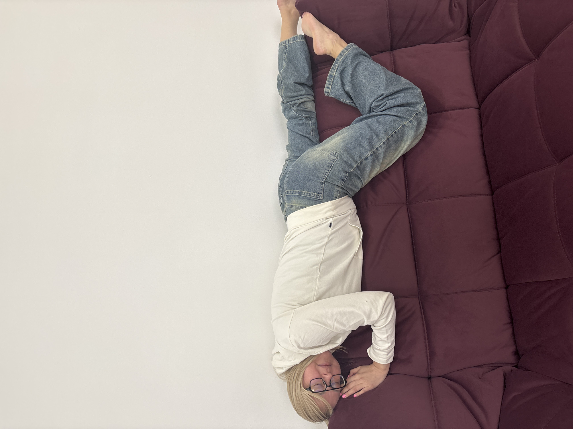 A woman lies upside down on a burgundy sofa. She is wearing glasses, a long-sleeved white shirt, and blue jeans. Her feet point upwards towards a white wall. Her head rests on the burgundy sofa. The location is indeterminable without additional information beyond the image. The city of Izhevsk, Russia, is not deducible from this image.