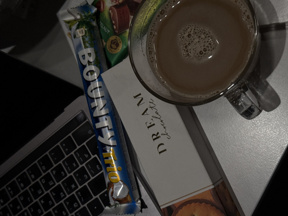 The image presents a static, overhead view of a desktop or table setup, dimly illuminated. In the upper left, the keyboard of a silver laptop is partially visible, displaying both Latin and Cyrillic characters. A blue and white 'Bounty Trio' chocolate bar wrapper lies diagonally across the top of the keyboard. Below this, a white rectangular box labeled "DREAM chocolate" in gold text is positioned. Adjacent to the chocolate box and partially obscured are several individually wrapped candies, including a green wrapper with text suggesting "HAZELNUT" content. A clear glass mug, filled with a frothy light brown liquid, occupies the lower right quadrant of the frame. No individuals or specific actions are depicted. The objects rest on a smooth, light-colored surface. While the laptop's Cyrillic keyboard layout is consistent with usage in the region including Armenia, the image lacks specific architectural features, landmarks, or identifying signage to pinpoint an exact location within the city of Yerevan, Armenia.
