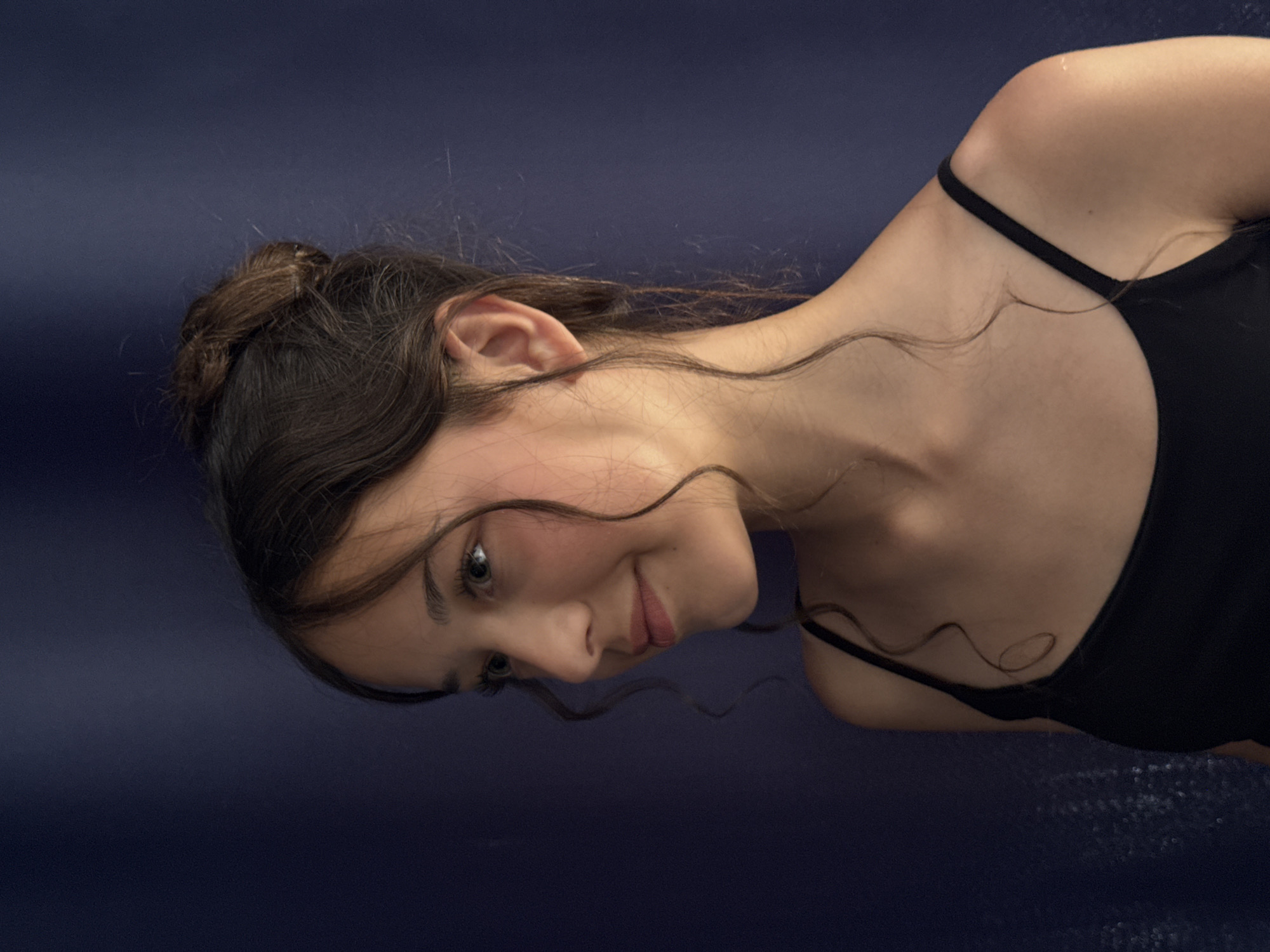 This is a close-up, vertically oriented portrait of a young woman. She has dark hair pulled back in a bun with wisps falling around her face and shoulders. She is looking downwards and to her left with a slight smile. Her skin appears fair, and she is wearing a black tank top with thin straps. The background is a dark, textured blue, suggesting water or a dark fabric. The lighting is soft, highlighting her features and creating a gentle mood. There are no visible text elements in the image. The scene is intimate and focused on the subject's expression and form, with a sense of calm or contemplation. Given the location context of Sochi, Russia, the dark blue background might suggest a pool or a body of water, possibly indicating leisure or athletic activity.